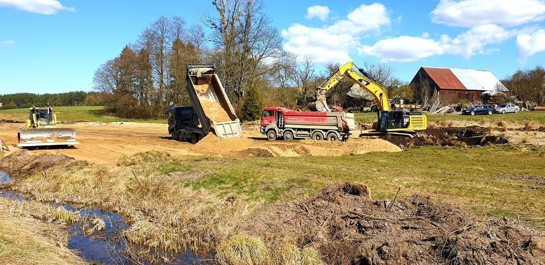Żółta koparka CAT ładuje ziemię na czerwoną ciężarówkę na budowie, w tle widoczny wywrotka z podniesioną skrzynią i spływającą ziemią, oraz spychacz i budynek gospodarczy z zaparkowanymi samochodami.