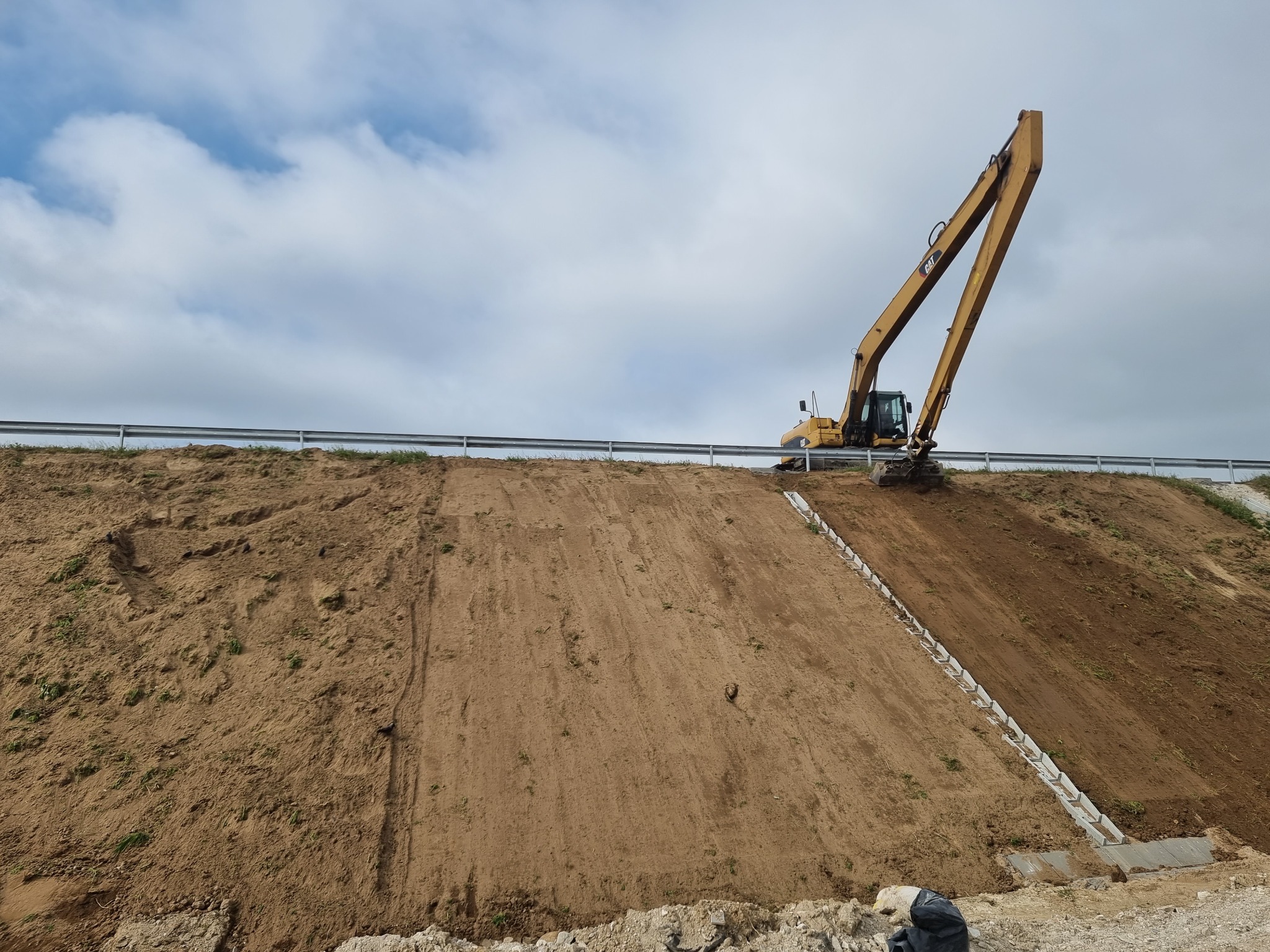 Żółta koparka na szczycie wzgórza z zabezpieczeniem, widoczne ślady pracy ziemnej i elementy betonowe na zboczu.
