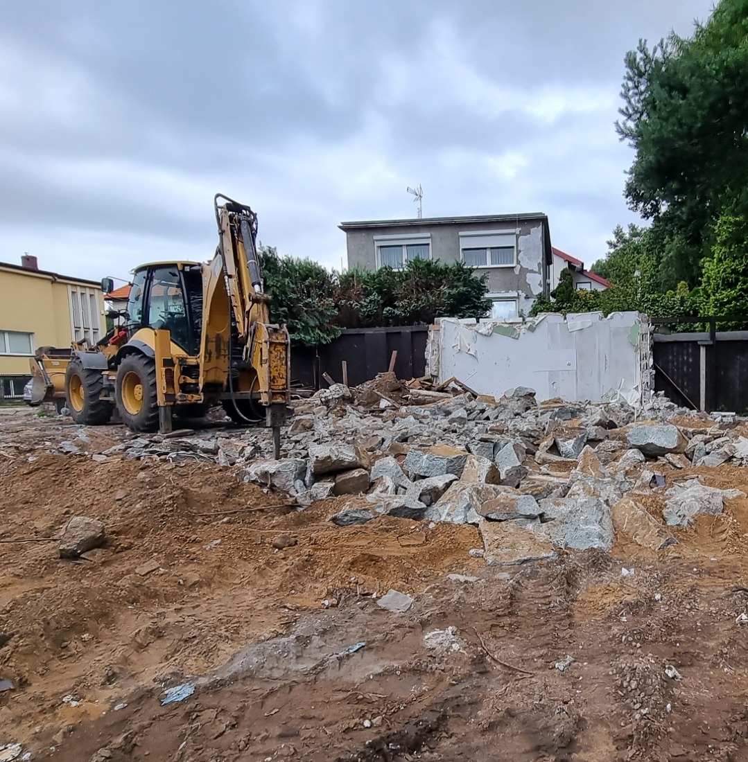 Żółta koparko-ładowarka z wiertłem hydraulicznym rozbija gruz na placu budowy, w tle budynki mieszkalne i pochmurne niebo.