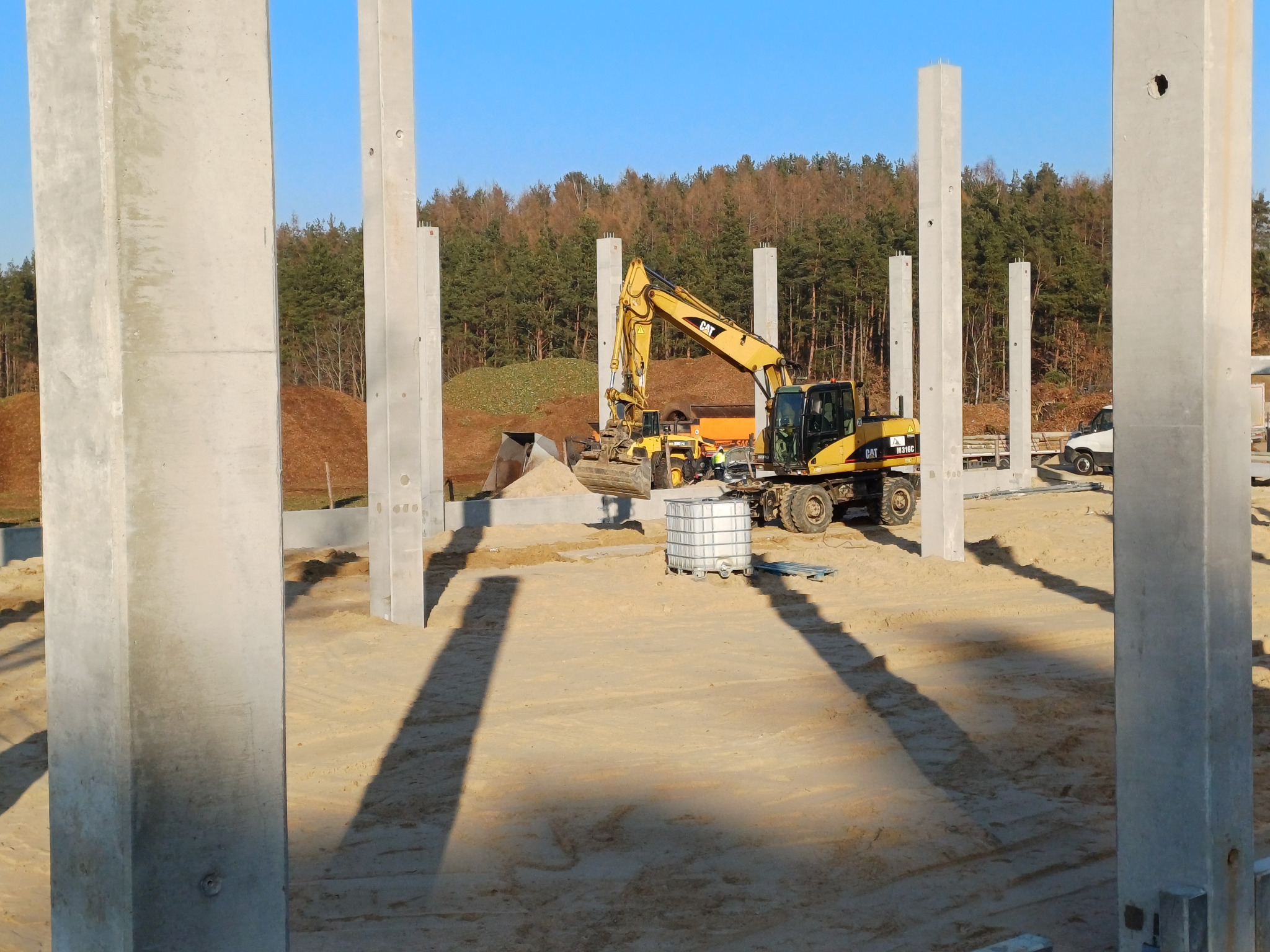 Żółta koparka przy wykopie fundamentów, widoczny szkielet konstrukcji z betonowych słupów, w tle las i błękitne niebo.