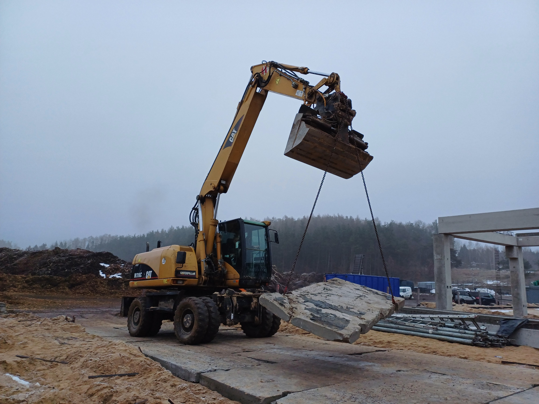 Żółta koparka CAT M316C podnosi betonowy element za pomocą łańcuchów na placu budowy, w tle widoczny las i konstrukcja budynku w surowym stanie.