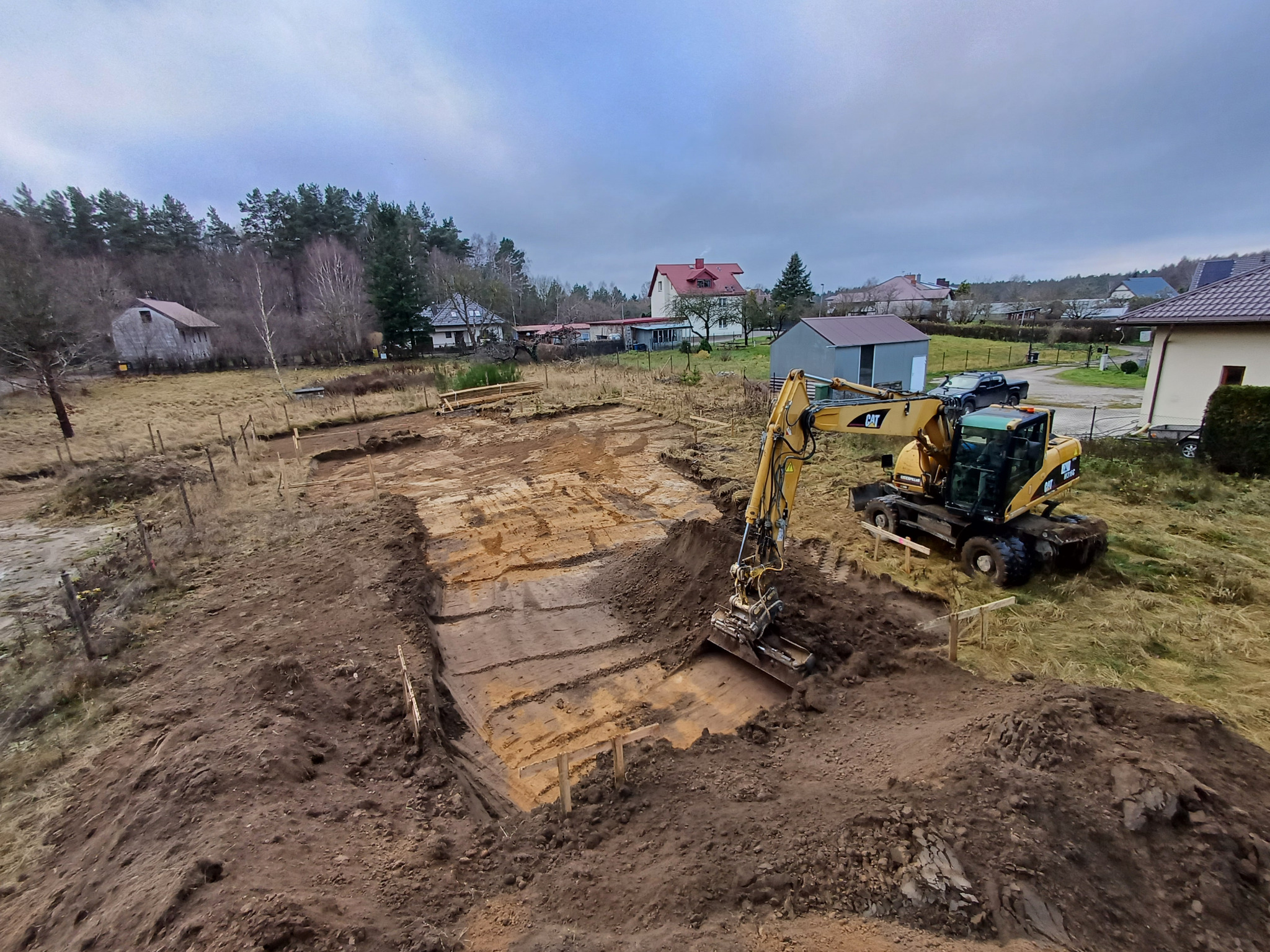 Żółta koparka CAT wykonuje wykop pod fundamenty na działce z widocznymi drewnianymi szalunkami, w tle zabudowa wiejska pod pochmurnym niebem.