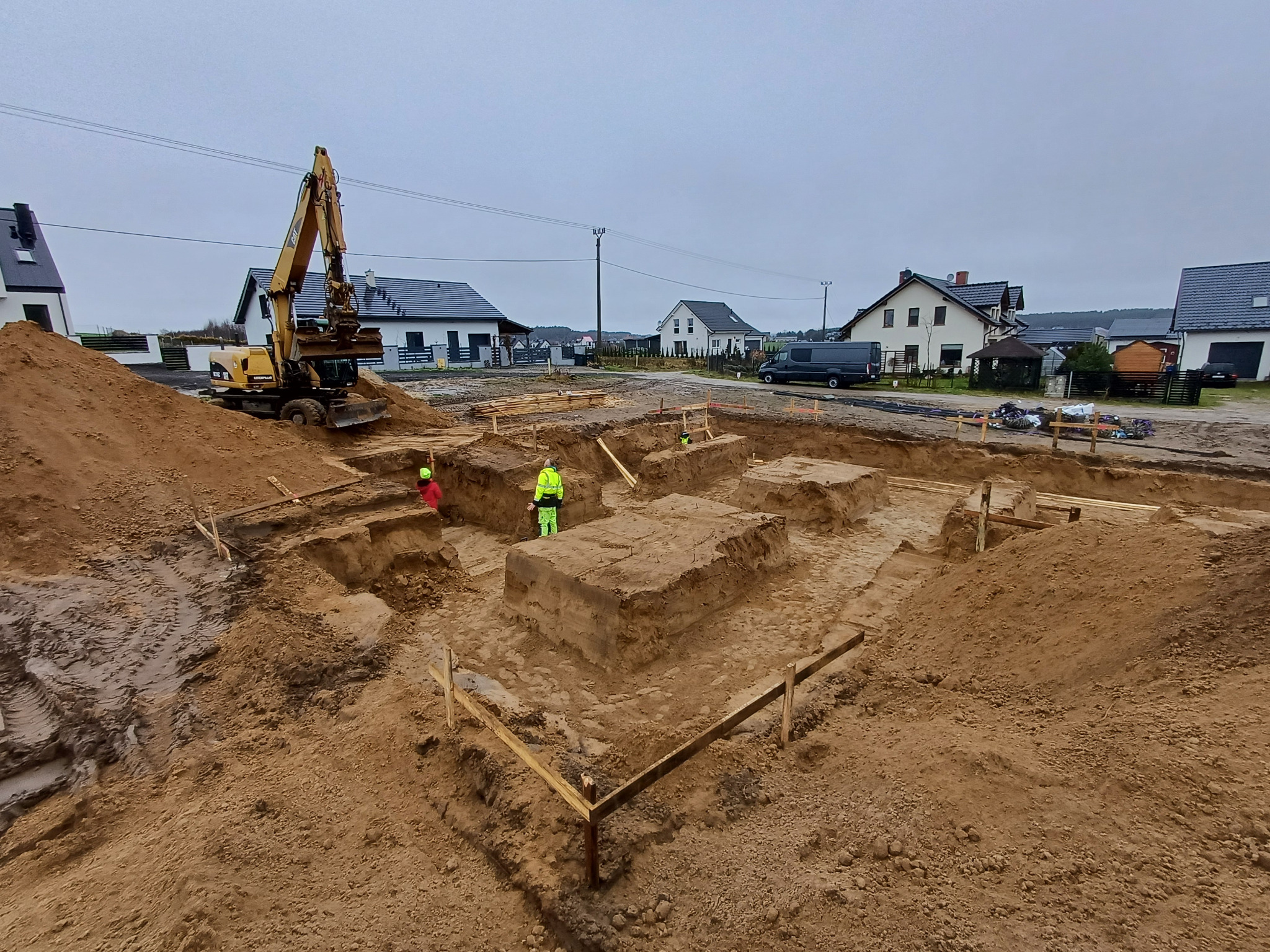 Wykop pod fundamenty z widocznymi ławami fundamentowymi, szalunkami, pracującą koparką i pracownikami w odblaskowych ubraniach na tle zabudowy jednorodzinnej w pochmurny dzień.