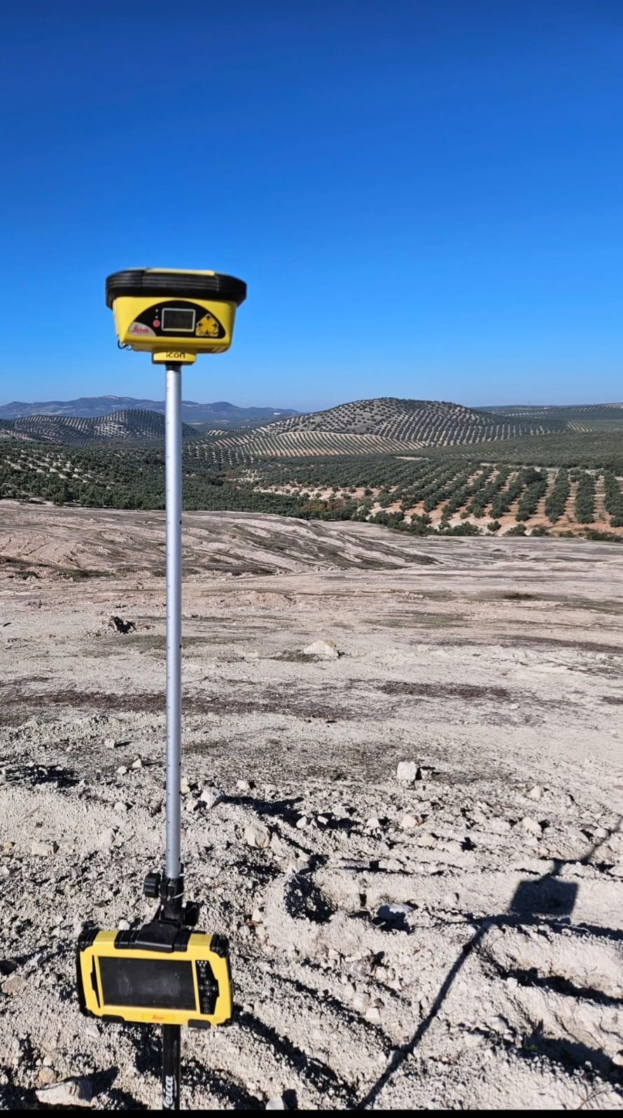 Żółty odbiornik GNSS Leica Icon na statywie, na nierównym terenie z widokiem na wzgórza obsadzone drzewami w tle, pod błękitnym niebem.