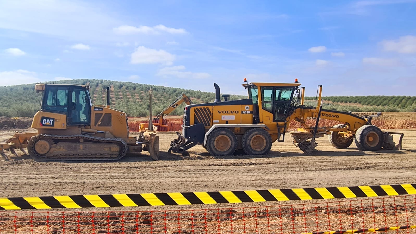 Żółty spychacz CAT i równiarka VOLVO G940 na wyrównanym terenie, z widoczną taśmą ostrzegawczą i drzewami w tle pod błękitnym niebem.