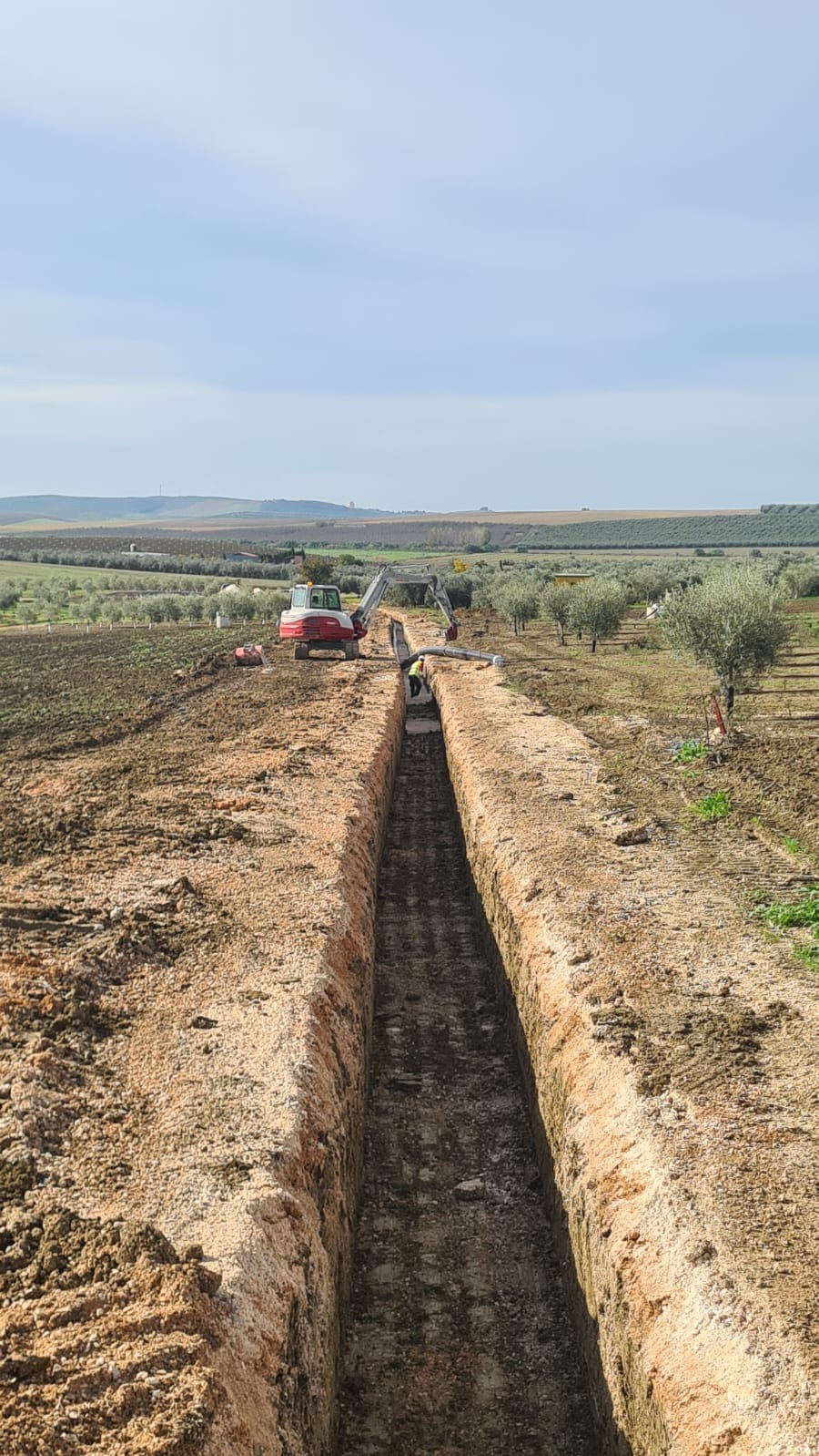 Długi, wąski wykop w ziemi, na jego końcu pracownik i koparka. W tle pola i drzewa pod błękitnym niebem. Wykop świeżo wykonany, o regularnych ścianach.