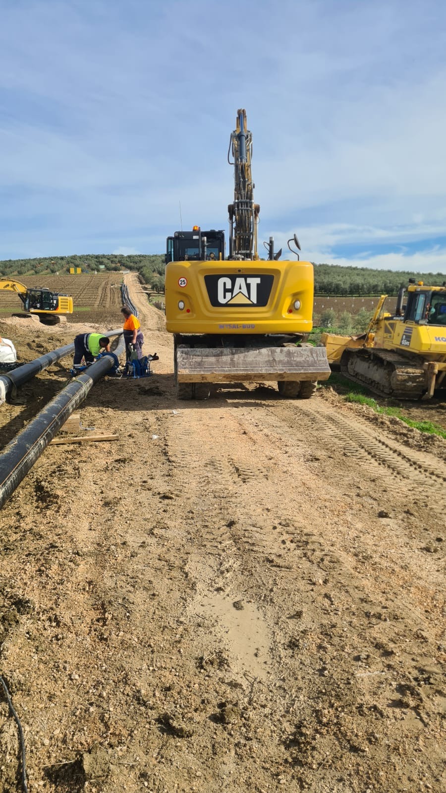 Żółta koparka CAT na placu budowy, w tle wzgórza i ekipa montująca rury. Widoczne ślady opon na ziemi i inne maszyny. Prace ziemne w toku.