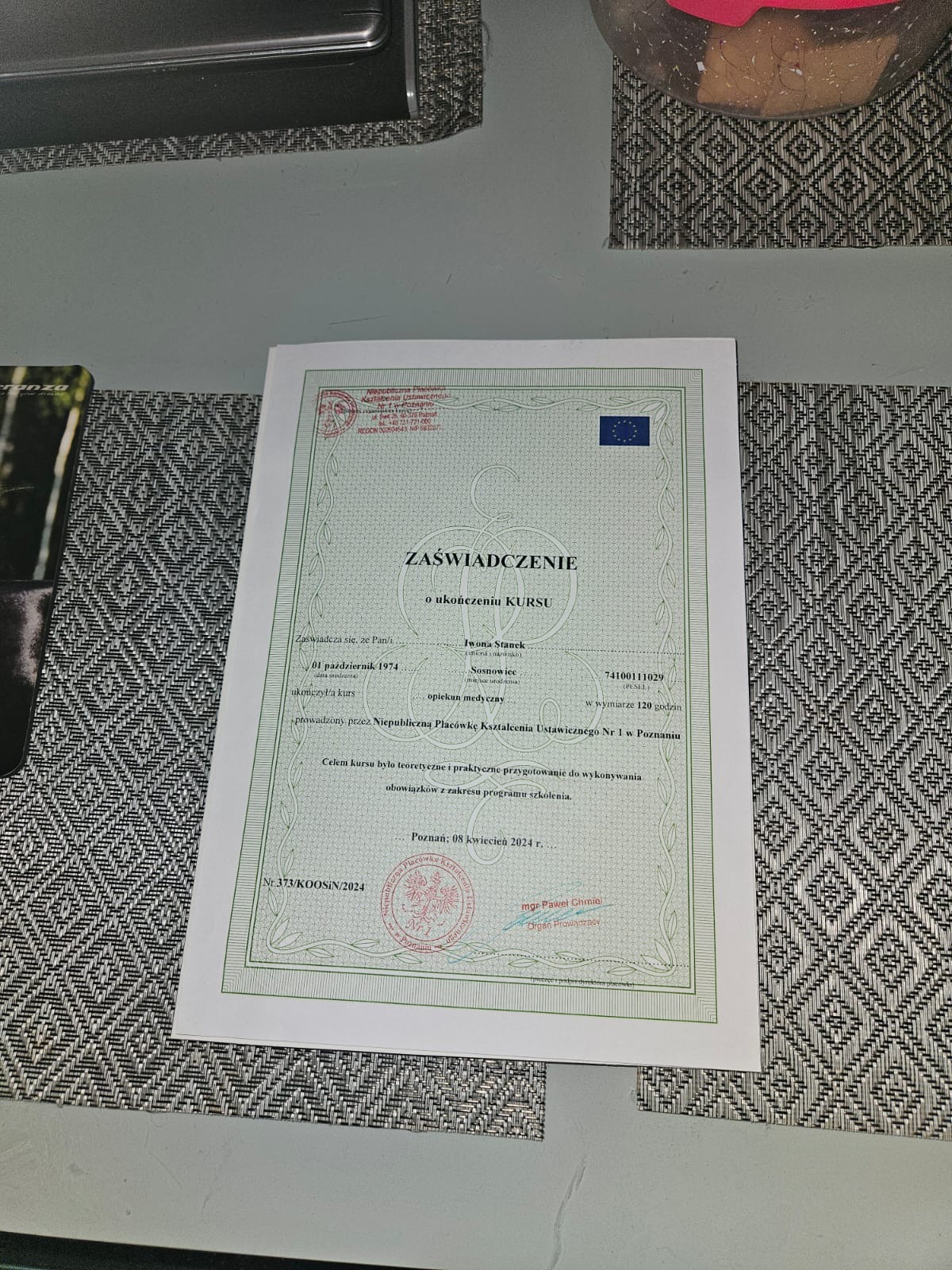 Skan zaświadczenia o ukończeniu kursu 'opiekun medyczny' przez Iwonę Stanek, wydanego w Poznaniu, z datą 08 kwietnia 2024 r., leżący na wzorzystej podkładce.