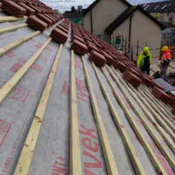 Układanie dach&oacute;wki ceramicznej na łatach z folią izolacyjną, widoczni dekarze w odblaskowych ubraniach, domy w tle.