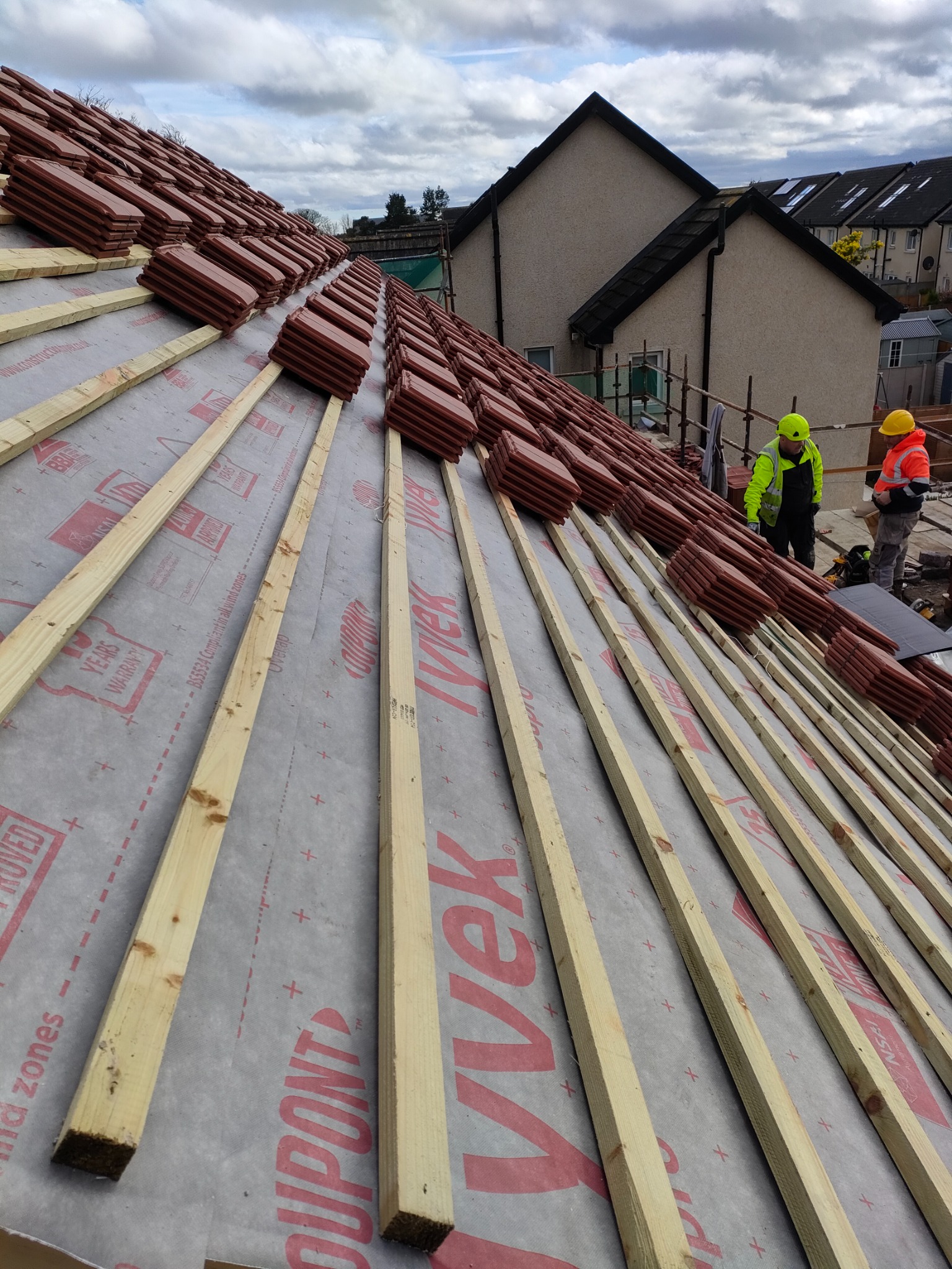 Układanie dachówki ceramicznej na łatach z folią izolacyjną, widoczni dekarze w odblaskowych ubraniach, domy w tle.