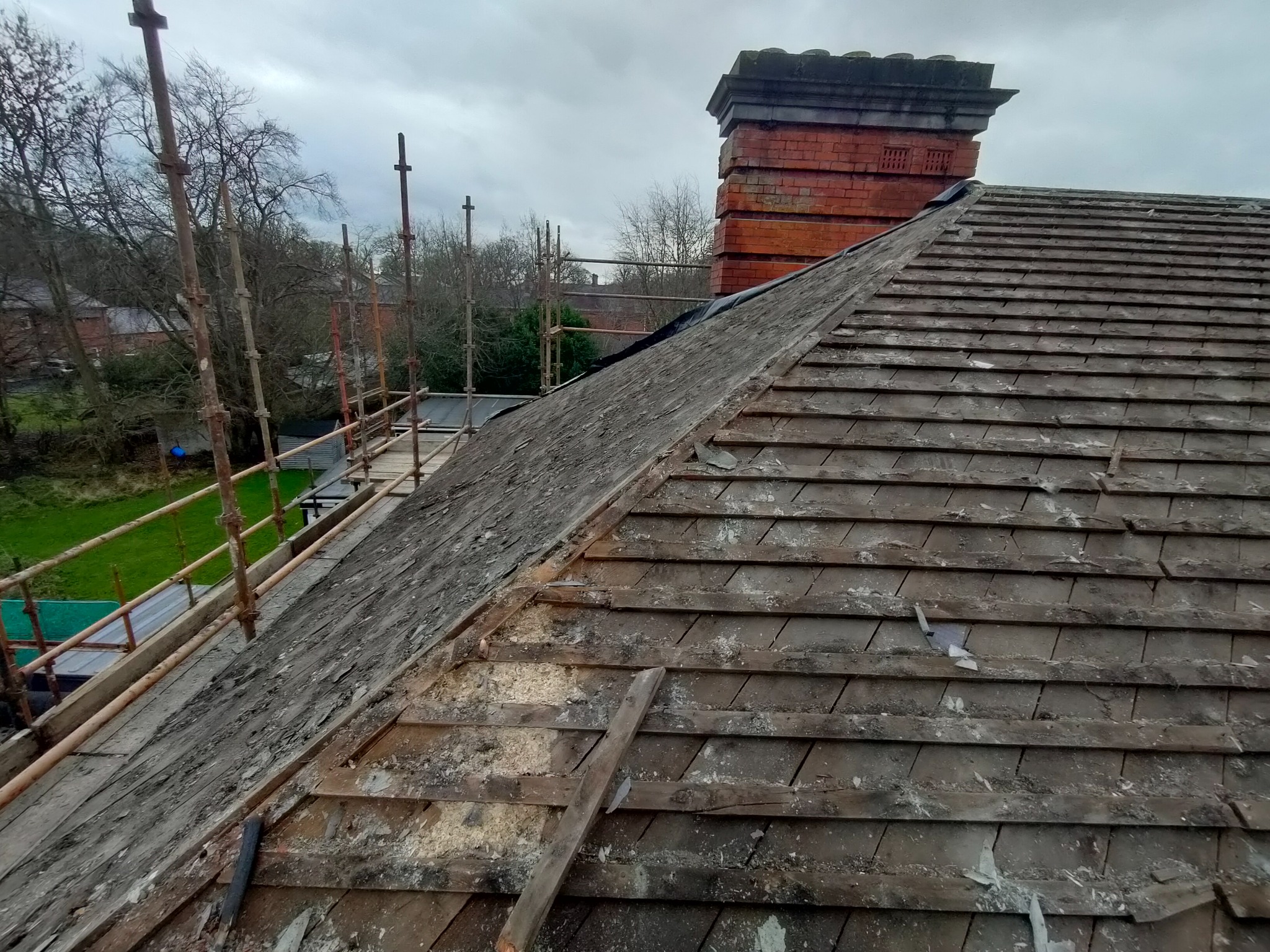 Demontaż starego pokrycia dachowego z łupka, widoczne rusztowanie, komin z czerwonej cegły i fragmenty uszkodzonych dachówek.