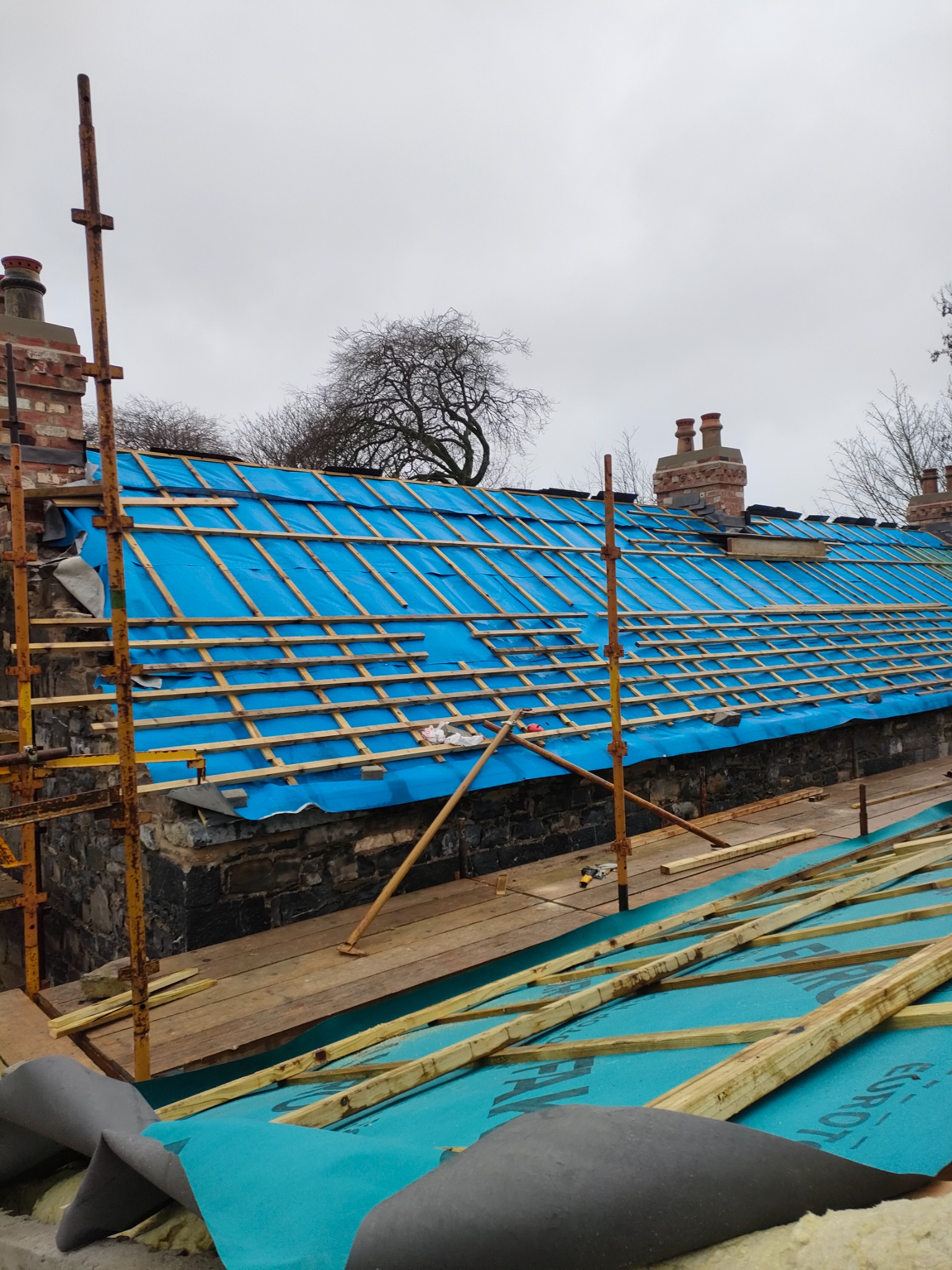 Dach budynku w trakcie renowacji, zabezpieczony niebieską membraną i drewnianą konstrukcją łat, widoczne kominy i rusztowanie.