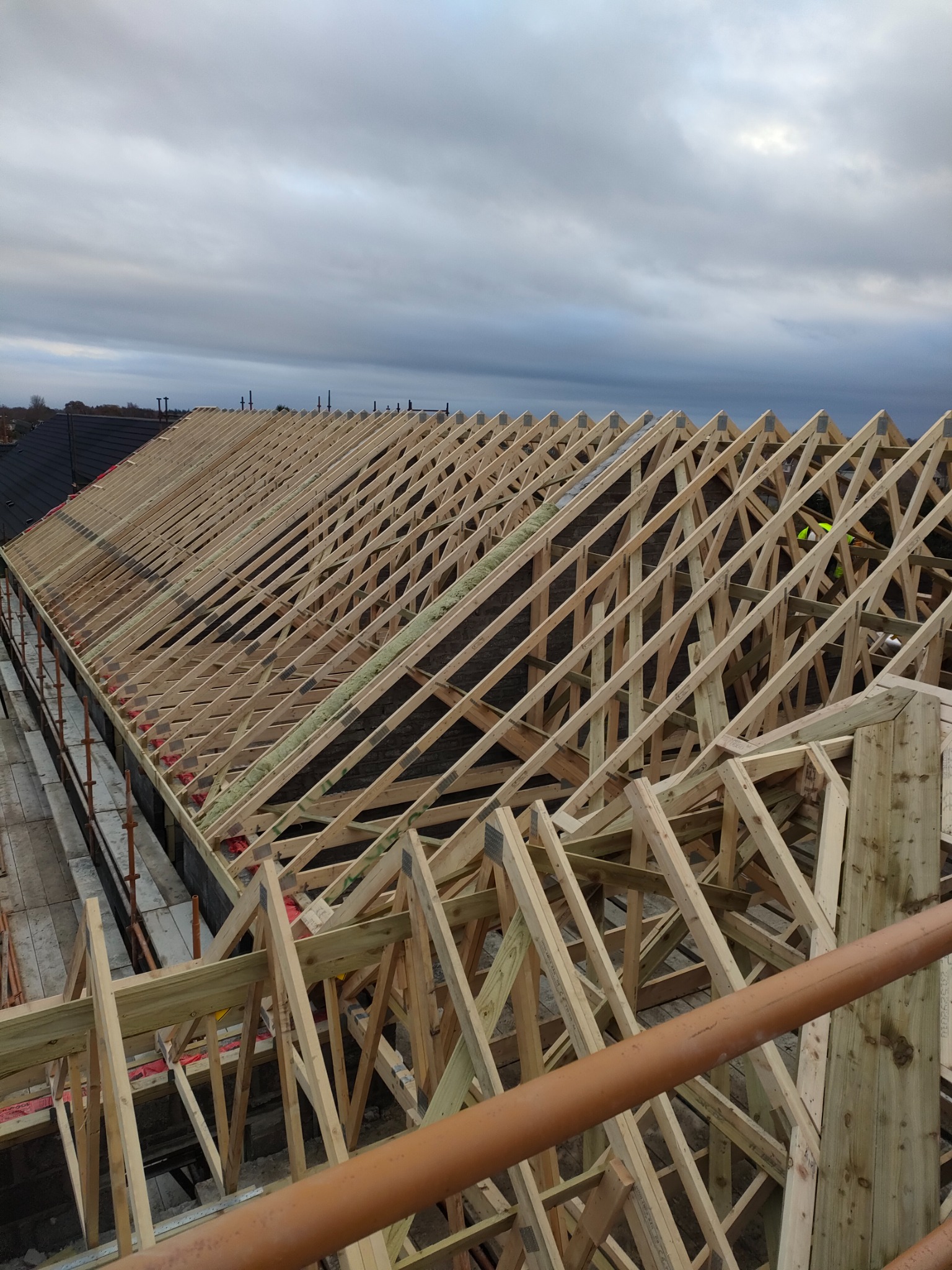 Wysoki kąt widzenia na konstrukcję drewnianą dachu w trakcie budowy, widoczne fragmenty izolacji, rusztowania i fragmenty budynku w tle, pochmurne niebo.