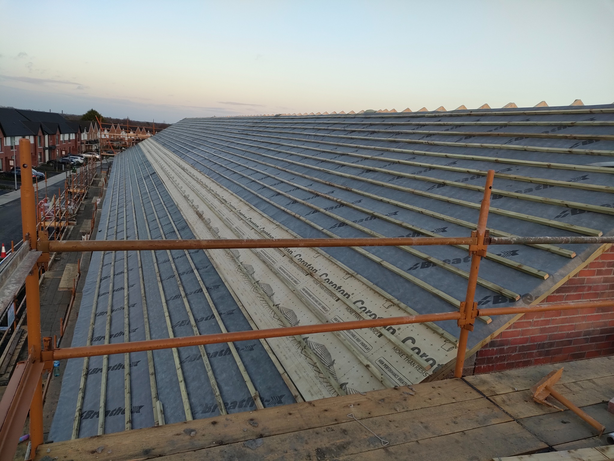 Konstrukcja dachu w trakcie budowy, widoczne membrany dachowe z nadrukiem, łaty drewniane, rusztowanie i fragment muru szczytowego. W tle zabudowa szeregowa i zachodzące słońce.