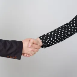 Uścisk dłoni pomiędzy mężczyzną w ciemnym garniturze z brązowymi guzikami a kobietą w czarnej sukience w białe grochy.