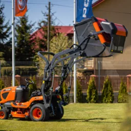 Pomarańczowy traktorek ogrodowy Kubota z podnośnikiem i koszem na trawę, stojący na zadbanym trawniku w słoneczny dzień.