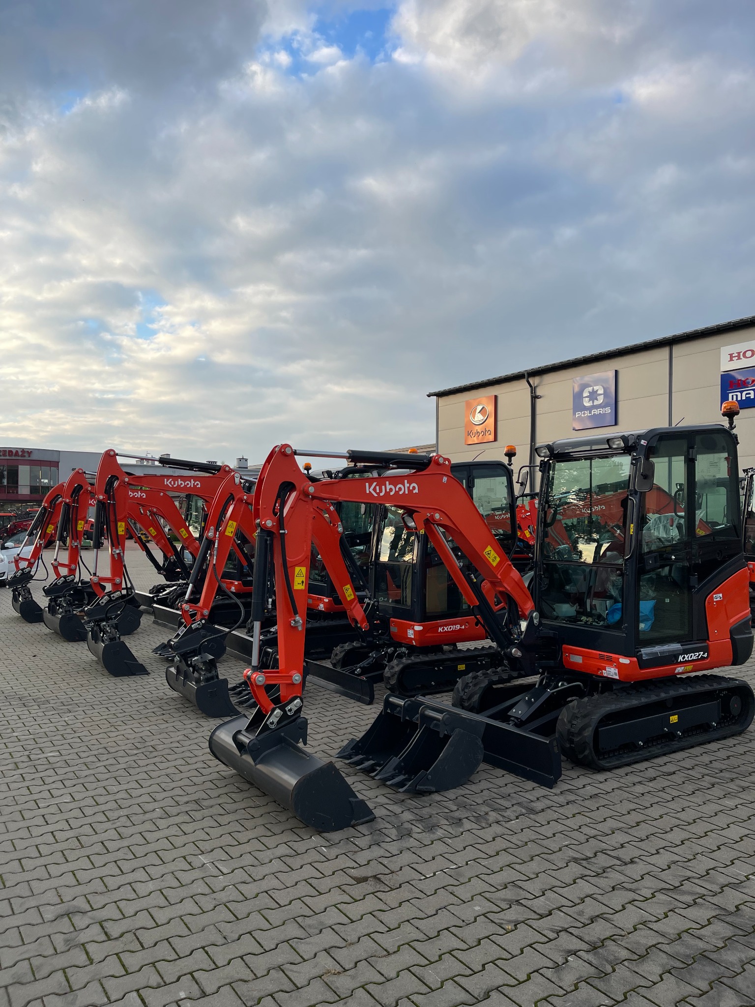 Rząd nowych, pomarańczowych minikoparek Kubota zaparkowanych na kostce brukowej przed budynkiem z logo Kubota i Polaris na elewacji, pochmurne niebo w tle.