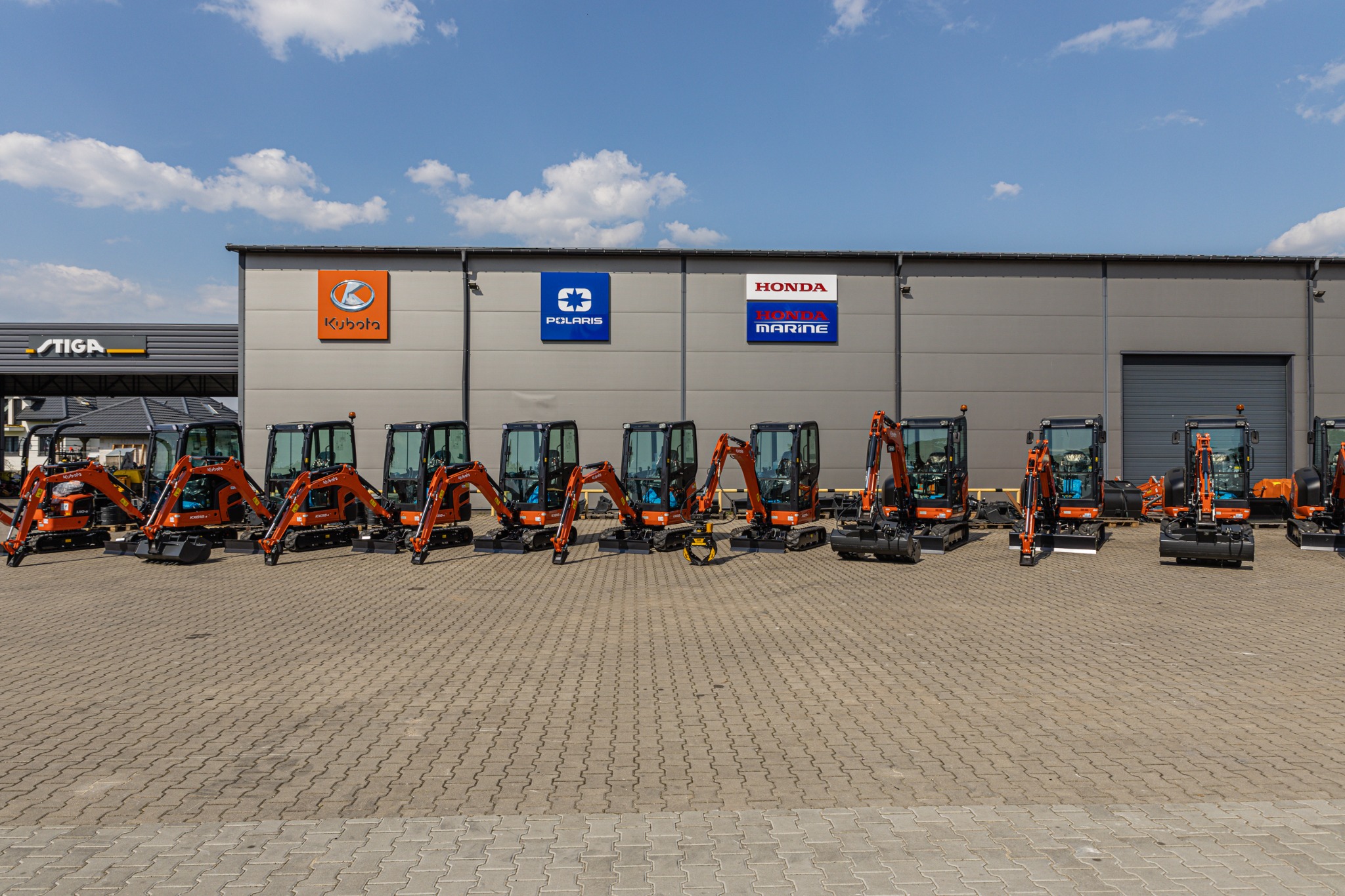 Rząd pomarańczowych maszyn budowlanych zaparkowanych przed szarym budynkiem z logotypami Kubota, Polaris i Honda Marine pod błękitnym niebem z chmurami.