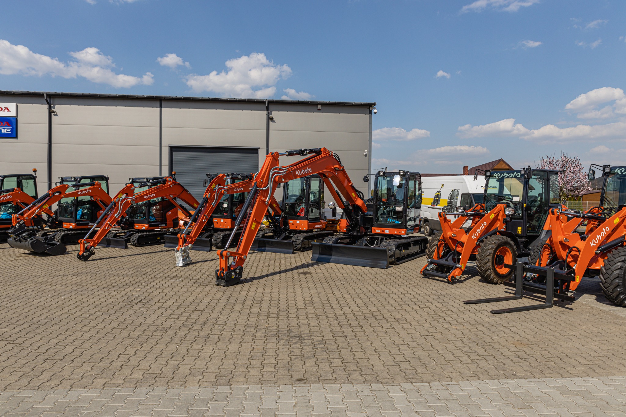 Rząd pomarańczowych maszyn budowlanych Kubota, w tym koparki i ładowarki, zaparkowanych na placu przed budynkiem z logo Honda, w słoneczny dzień.