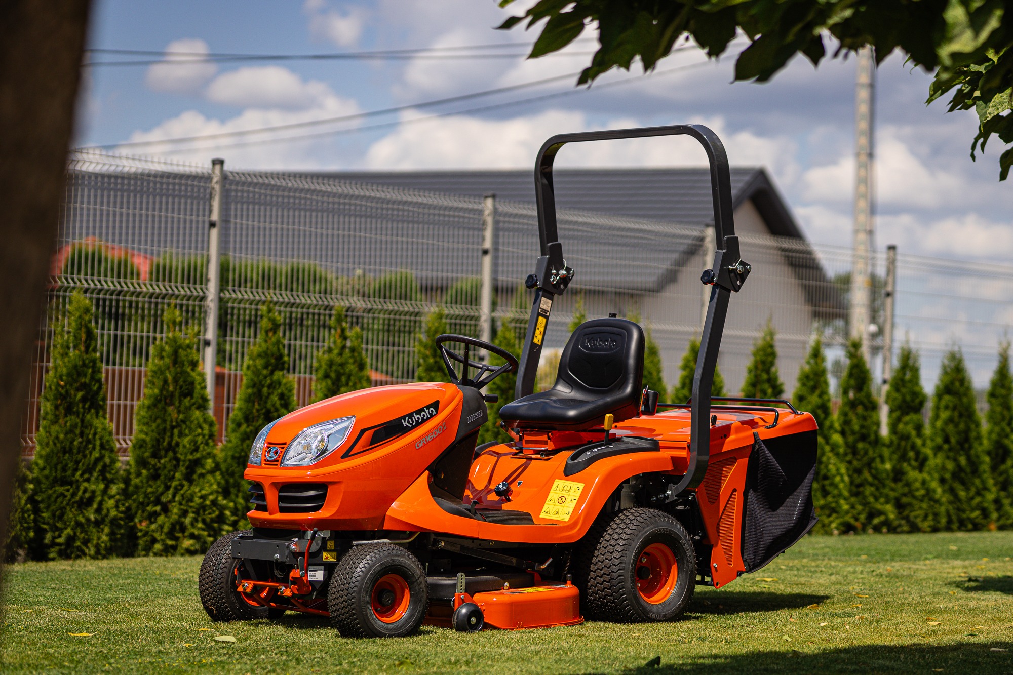 Pomarańczowy traktorek do koszenia trawy Kubota GR1600-I Diesel na trawniku przed ogrodzeniem z siatki i tujami w słoneczny dzień.