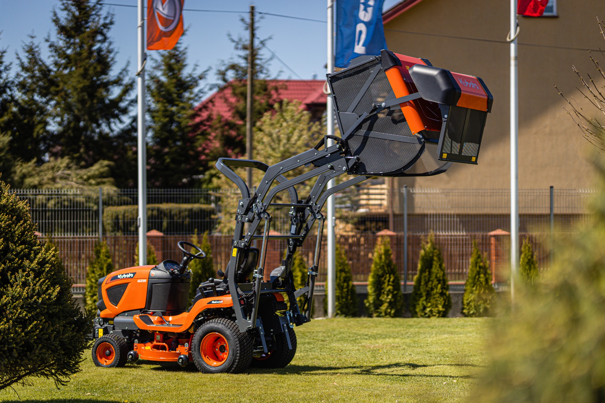 Pomarańczowy traktorek ogrodowy Kubota z podnośnikiem i koszem na trawę, stojący na zadbanym trawniku w słoneczny dzień.