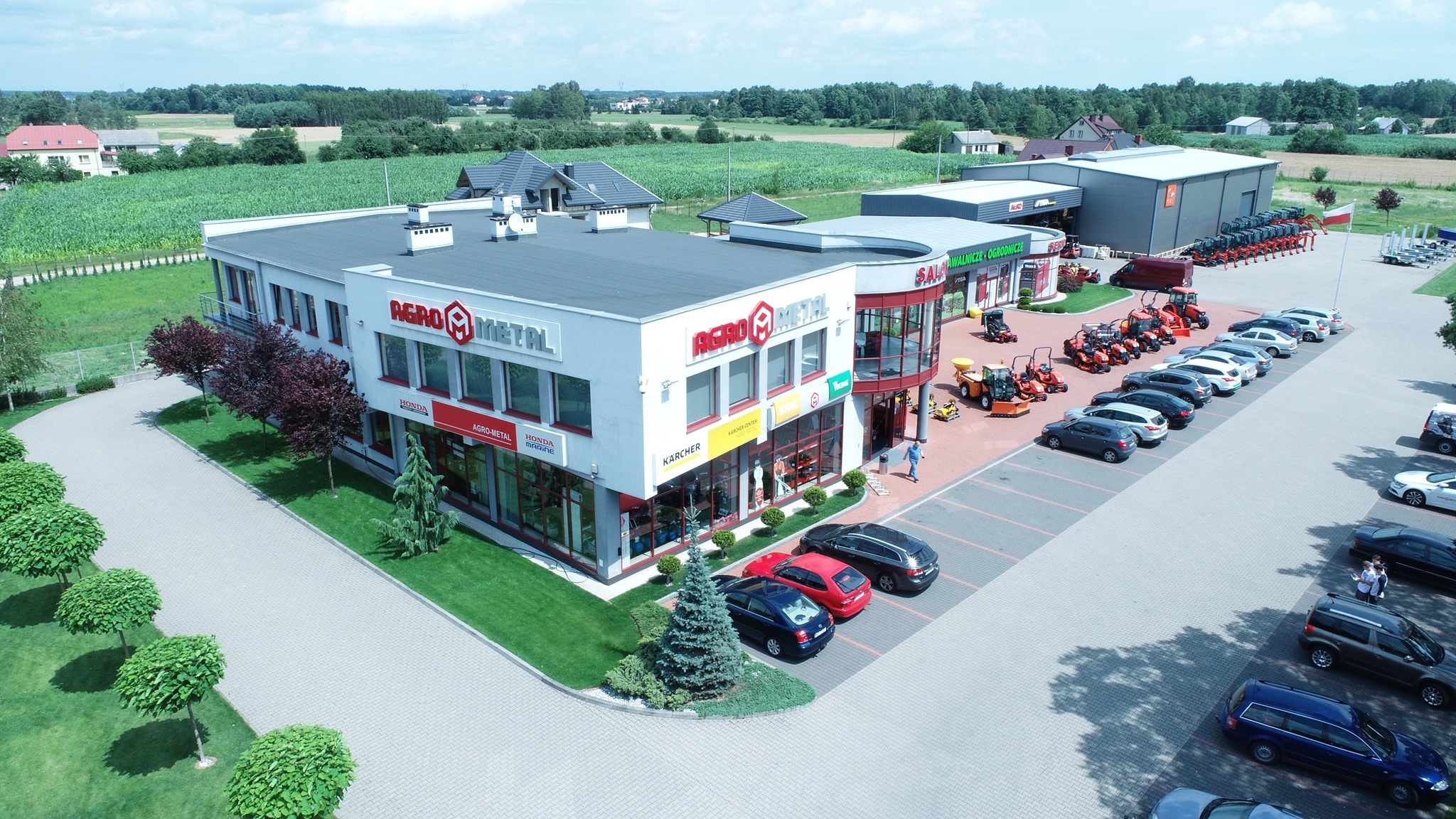 Widok z góry na salon sprzedaży maszyn rolniczych i ogrodniczych Agro-Metal z ekspozycją sprzętu przed budynkiem i na parkingu.