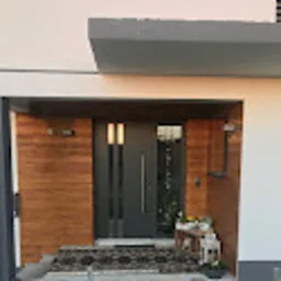 Nowoczesne, antracytowe drzwi wejściowe z dwoma pionowymi przeszkleniami, zamontowane w jasnej elewacji domu z drewnianą okładziną po bokach wejścia. Przed drzwiami wycieraczka i mały stolik...