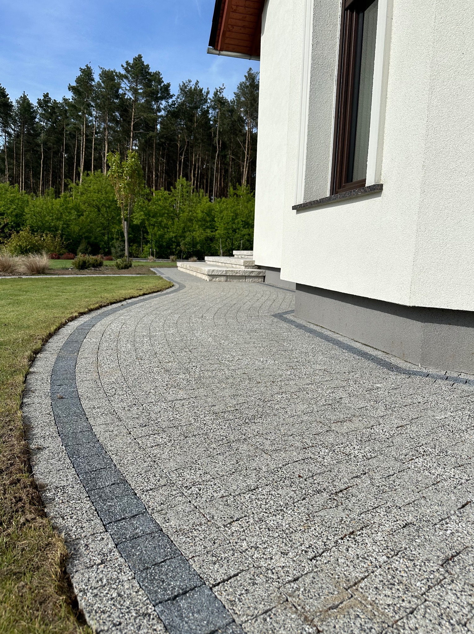 Kremowa kostka brukowa ułożona w łuk przy białej elewacji domu z ciemnobrązowym oknem, obrzeże z ciemnej kostki, trawnik i drzewa w tle.