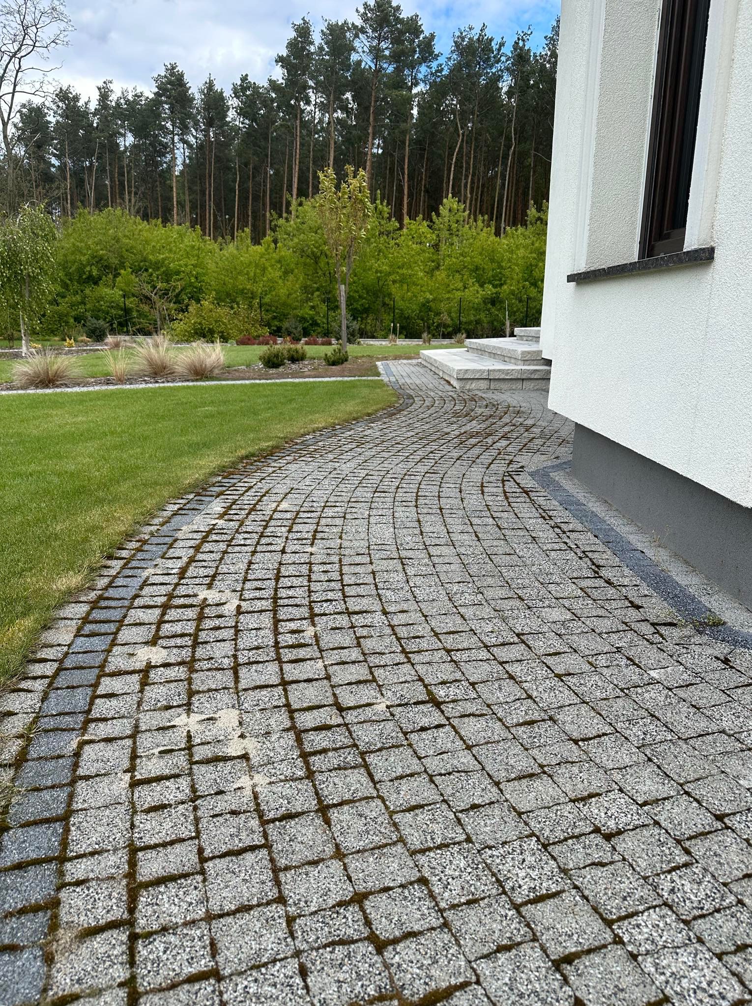 Szara kostka brukowa ułożona w łukowaty wzór prowadząca do schodów wejściowych budynku z białymi ścianami i ciemnymi oknami, otoczona zielonym trawnikiem i drzewami.