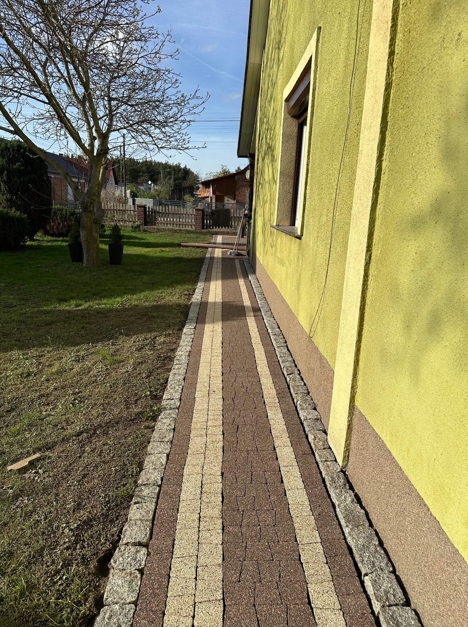 Ułożona ścieżka z kostki brukowej w dwóch kolorach, prowadząca wzdłuż ściany domu o żółtej elewacji, widok z perspektywy niskiego kąta.