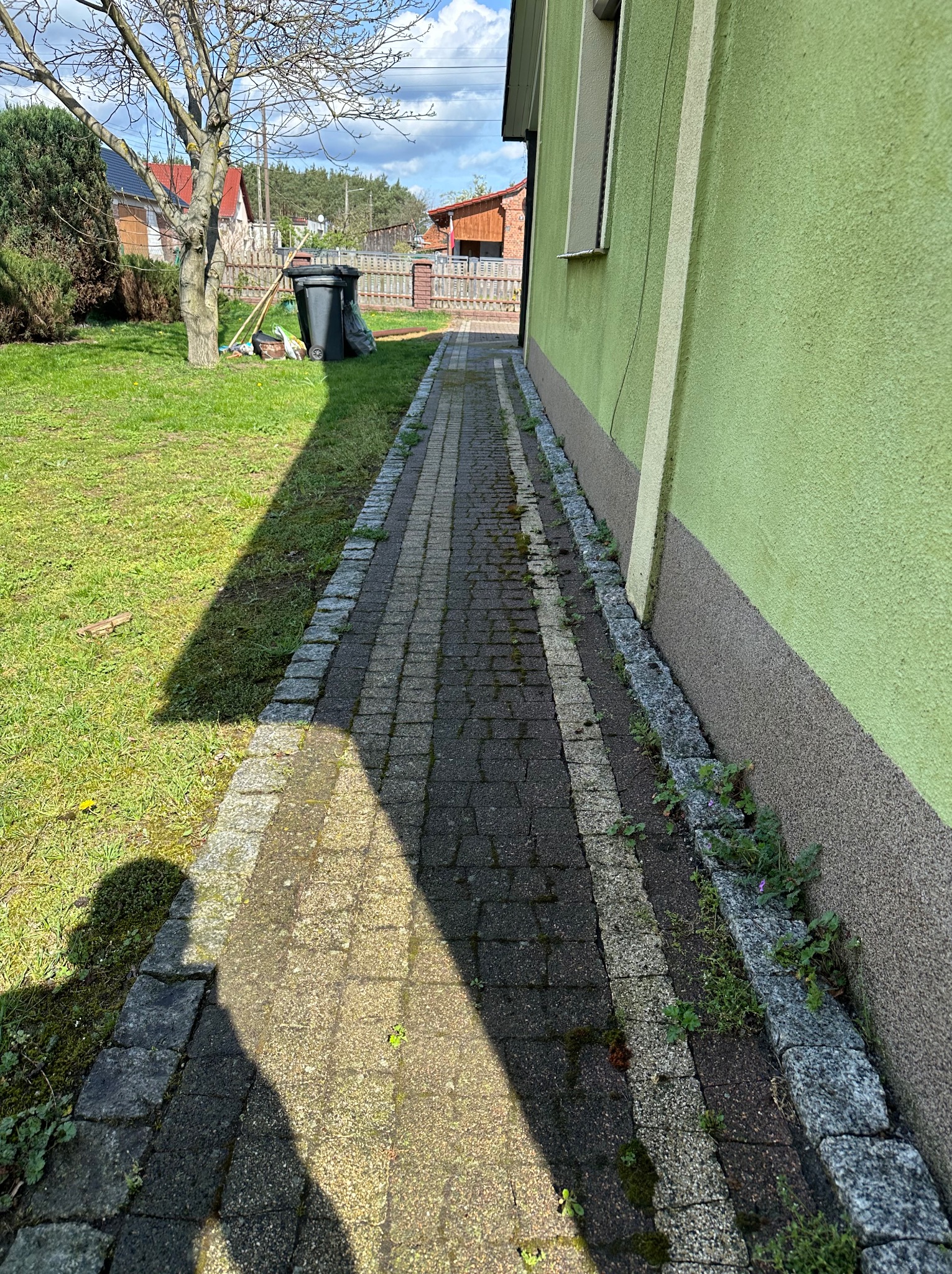 Chodnik z kostki brukowej prowadzący wzdłuż zielonej elewacji budynku, widoczne chwasty rosnące przy krawężniku i cień osoby fotografującej.
