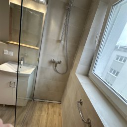 Gutak Project - Jasna łazienka z beżowymi płytkami, kabiną prysznicową ze szklaną ścianką, umywalką z szafką, oknem na poddaszu i uchwytem dla niepełnosprawnych przy oknie.