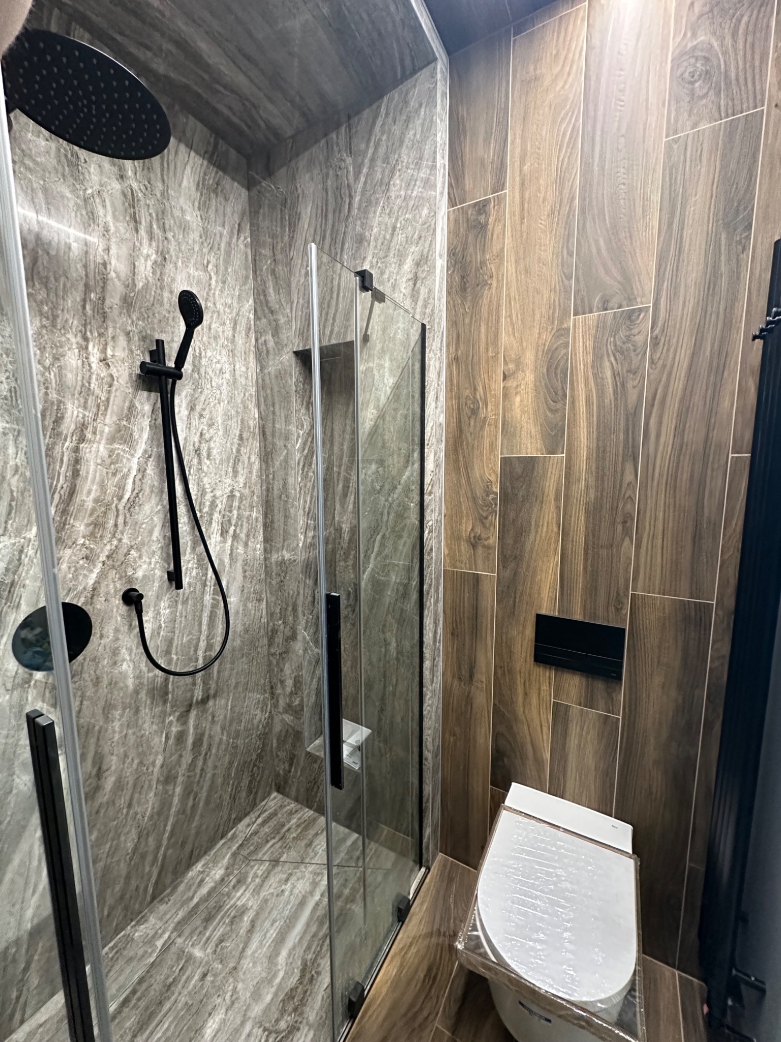 Nowoczesna kabina prysznicowa z czarną armaturą na tle marmurowych płytek, obok toaleta zabezpieczona folią ochronną i ściana wyłożona płytkami imitującymi drewno.