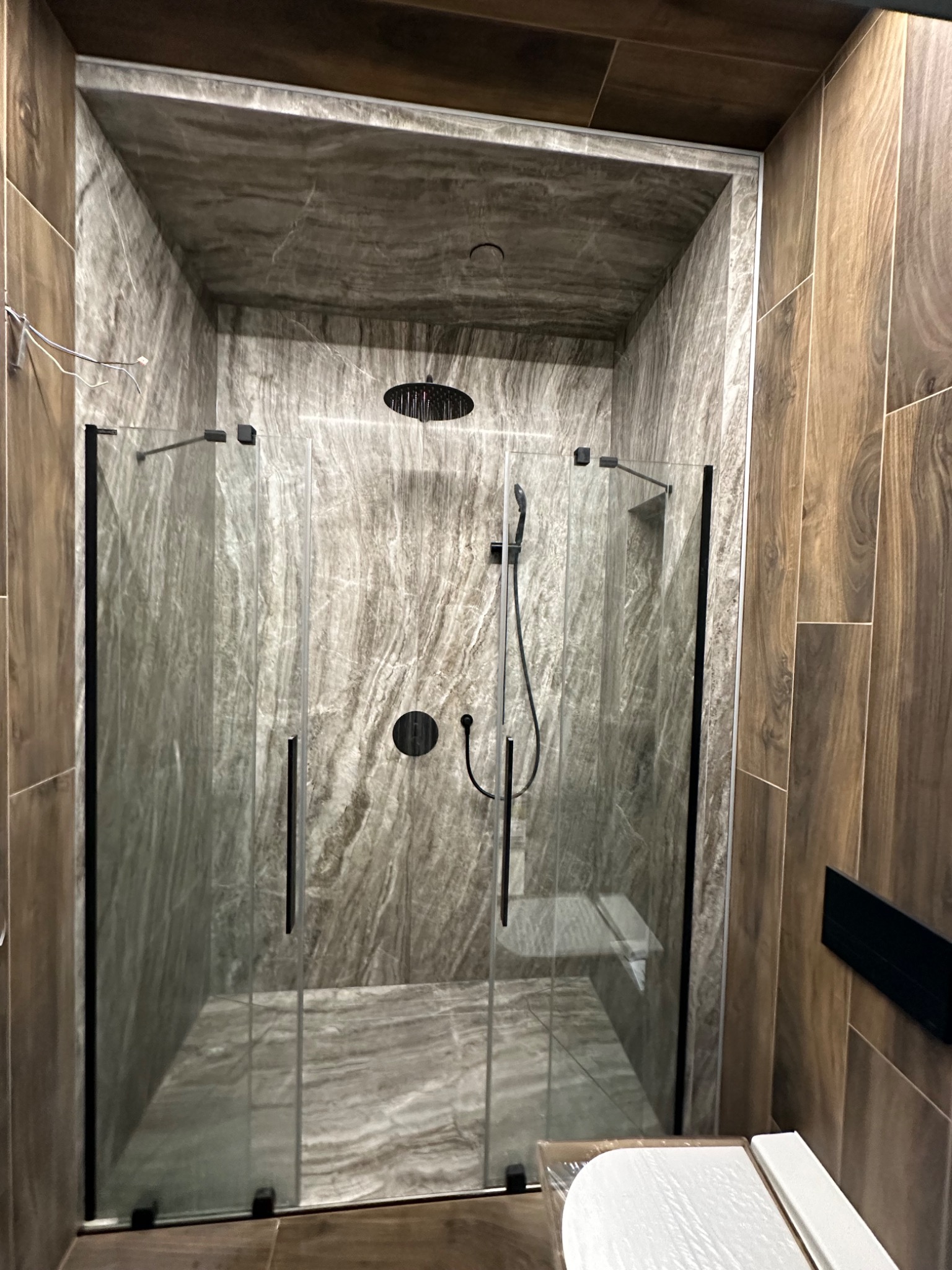 Kabina prysznicowa typu walk-in z czarnymi okuciami i deszczownicą, obłożona płytkami imitującymi marmur i drewno, widoczna z perspektywy wejścia do łazienki.