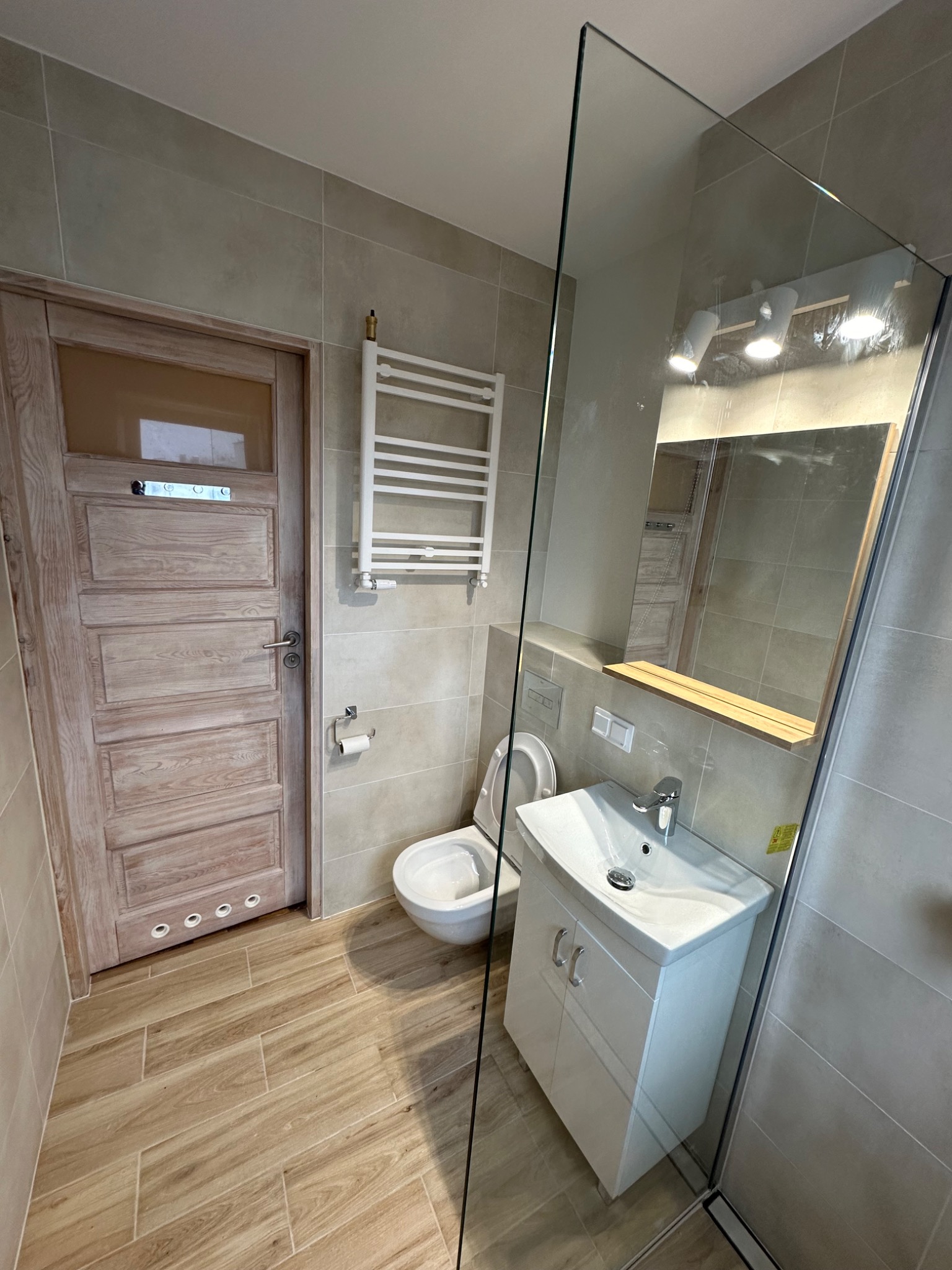 Jasna łazienka z drewnopodobnymi płytkami podłogowymi i beżowymi płytkami ściennymi, wyposażona w białą umywalkę z szafką, toaletę, grzejnik drabinkowy i kabinę prysznicową ze szkła.