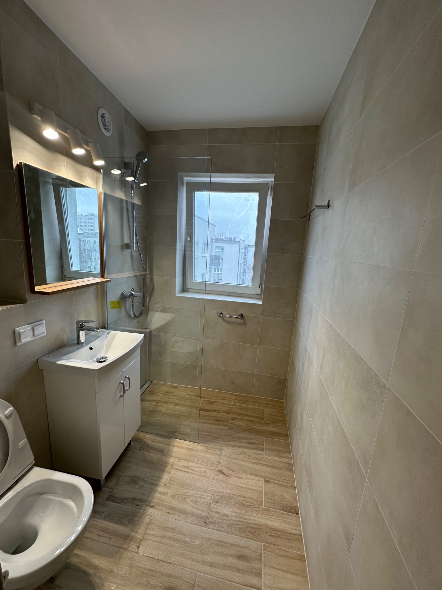 Nowoczesna łazienka z beżowymi płytkami imitującymi drewno na podłodze i ścianach, białą szafką pod umywalką, lustrem z oświetleniem punktowym, kabiną prysznicową ze szklaną ścianką oraz oknem...