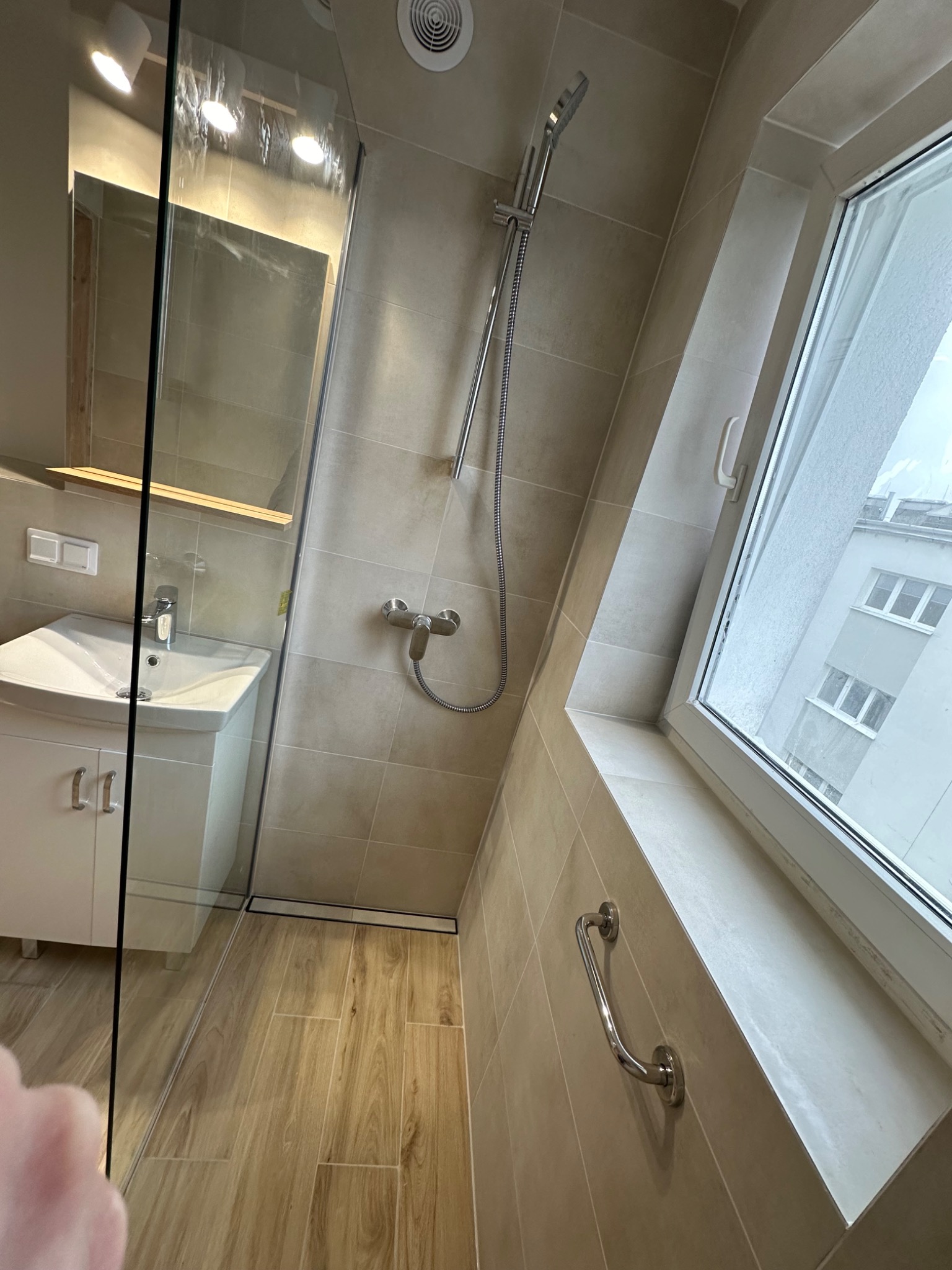 Jasna łazienka z beżowymi płytkami, kabiną prysznicową ze szklaną ścianką, umywalką z szafką, oknem na poddaszu i uchwytem dla niepełnosprawnych przy oknie.