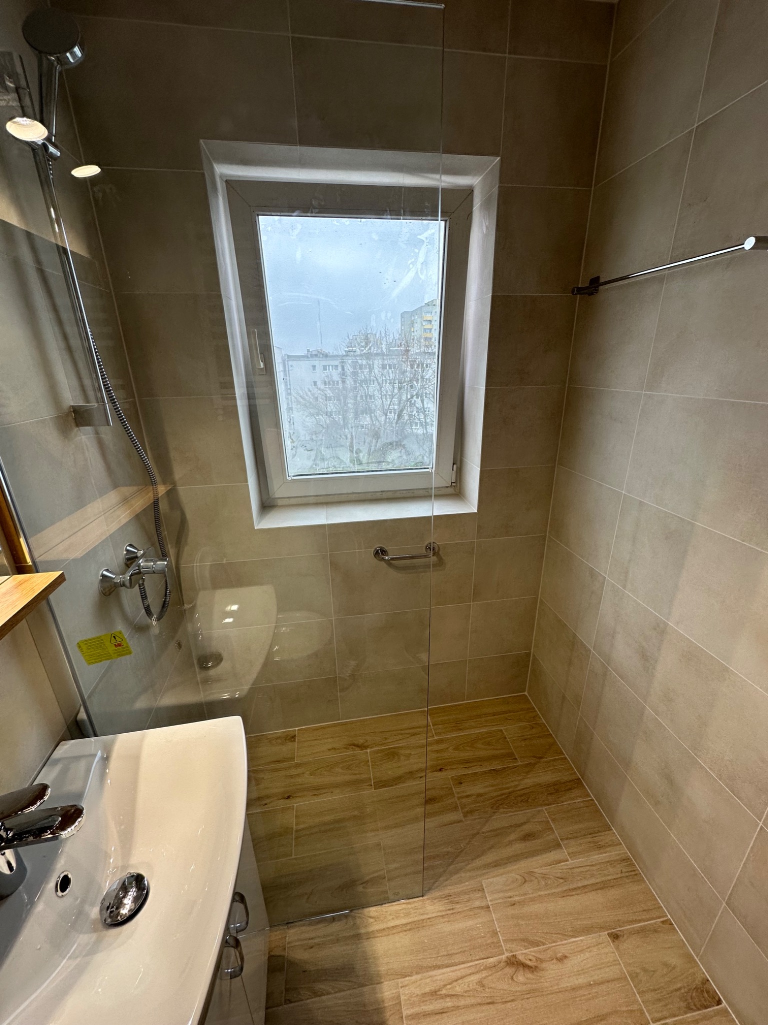 Nowoczesna łazienka z beżowymi płytkami, kabiną prysznicową ze szkła hartowanego i oknem z widokiem na budynki mieszkalne.