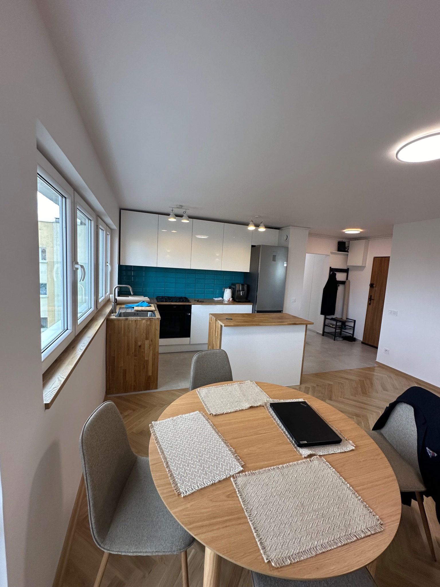 Nowoczesna kuchnia z białymi szafkami, turkusową glazurą i drewnianym blatem, widok z jadalni z okrągłym stołem i szarymi krzesłami, herringbone parkiet na podłodze.