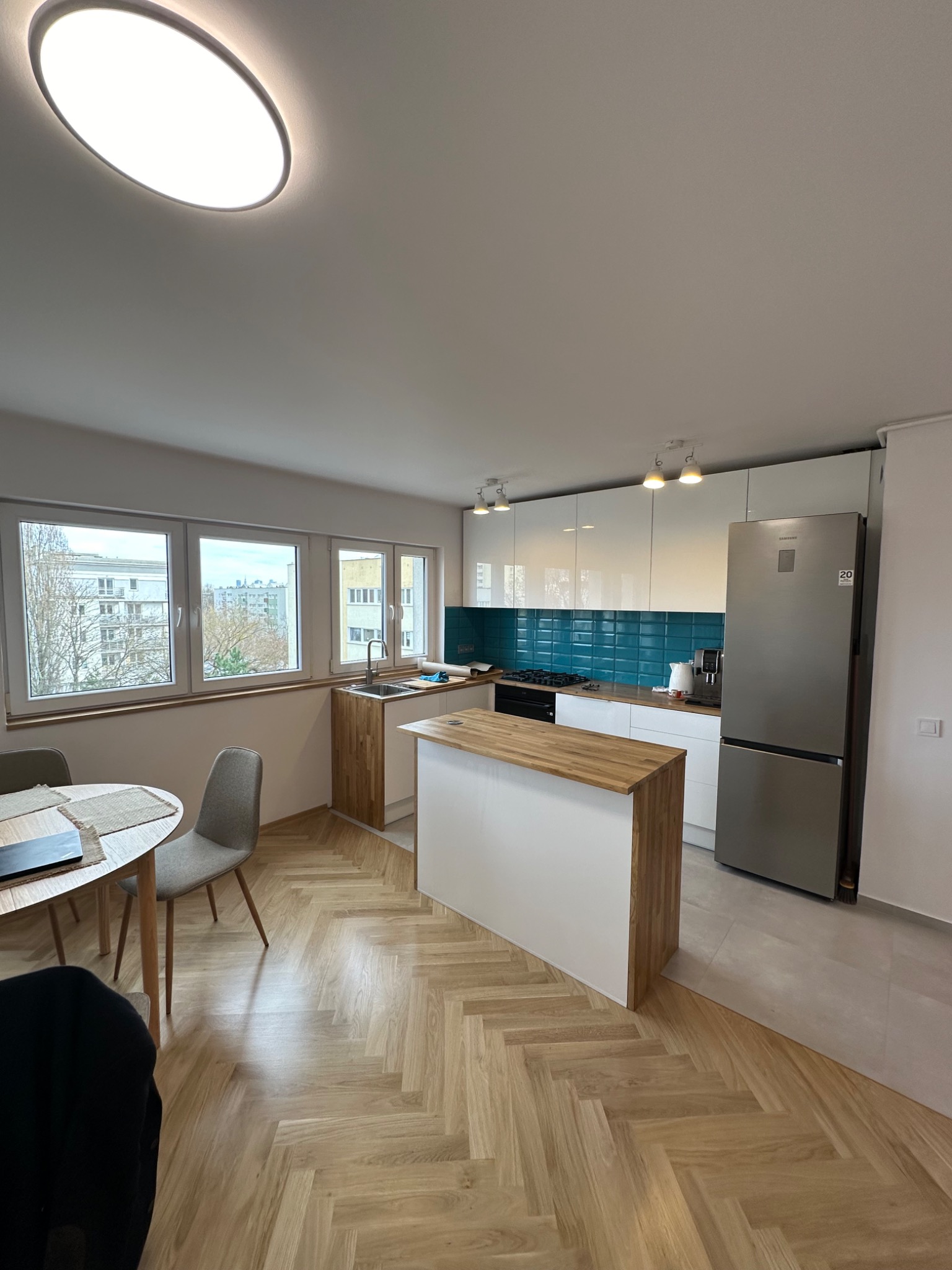 Jasna kuchnia z drewnianym blatem wyspowym i turkusowymi płytkami nad blatem, widok na stół z krzesłami i okno z widokiem na miasto.