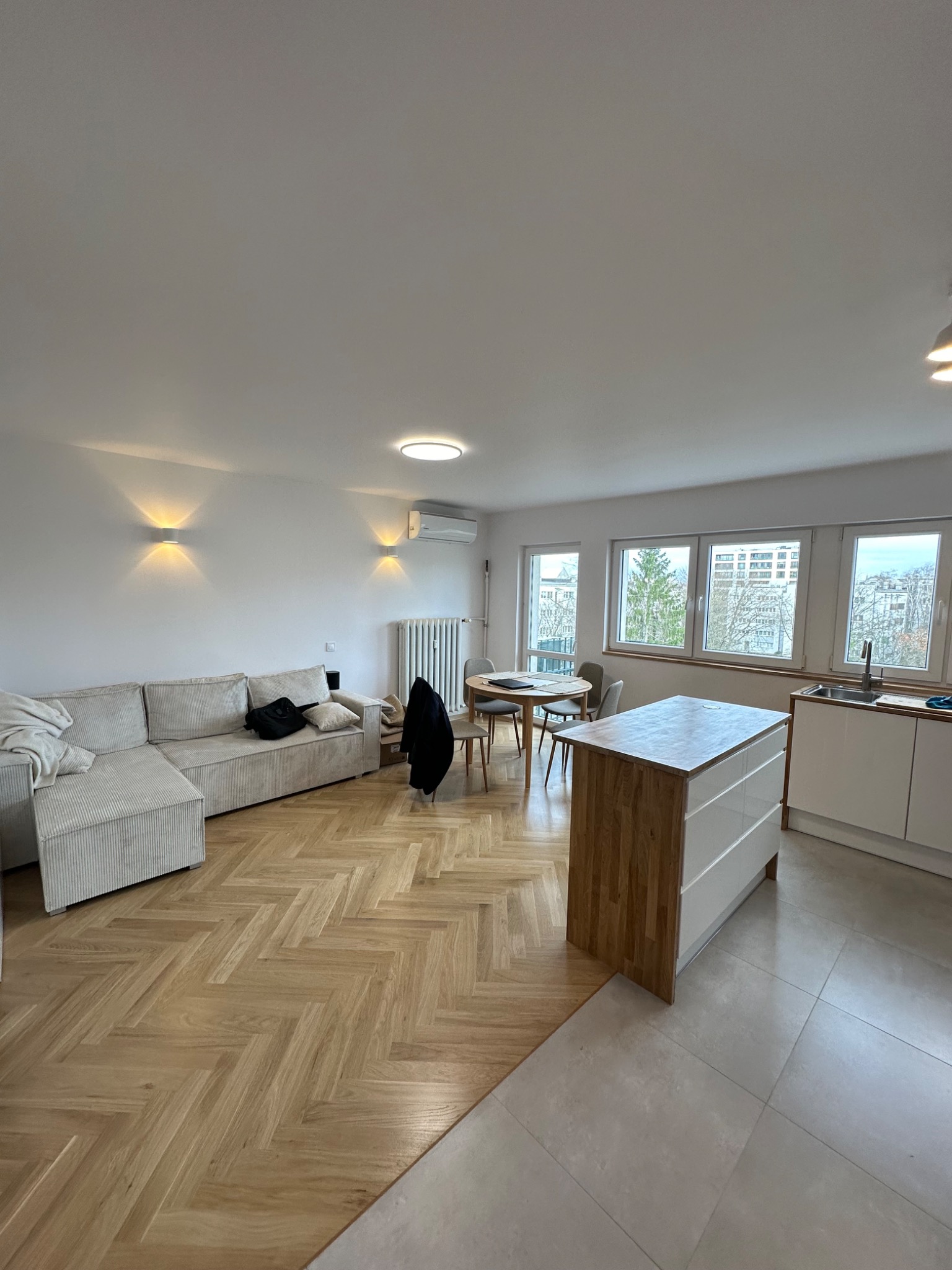 Jasne wnętrze mieszkania z parkietem w jodełkę, beżową sofą narożną, okrągłym stołem jadalnym i wyspą kuchenną z drewnianym blatem oraz białymi frontami szuflad.