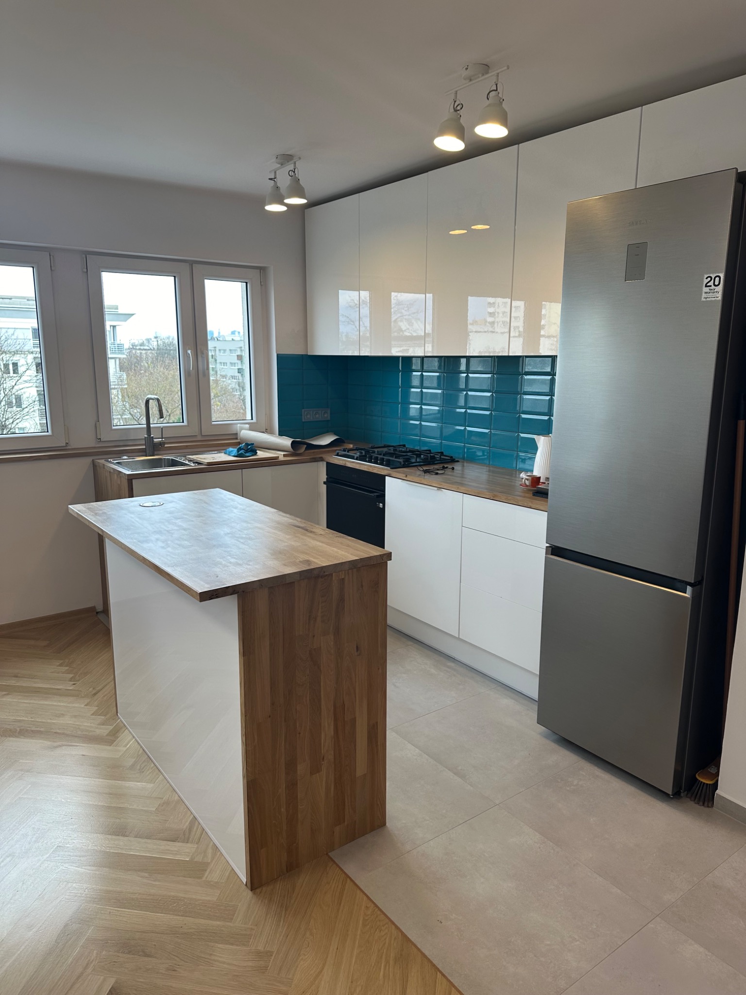 Nowoczesna kuchnia z wyspą, białe szafki, turkusowe płytki na ścianie, drewniany blat wyspy i parkiet w jodełkę, widok na miasto przez okno.
