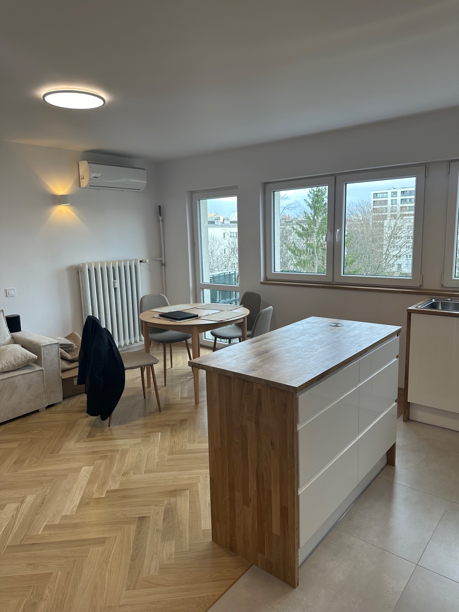 Jasne, przestronne wnętrze mieszkania z parkietem w jodełkę, wyspą kuchenną z drewnianym blatem i białymi szafkami, okrągłym stołem z krzesłami oraz grzejnikiem pod oknem z klimatyzatorem na ścianie.