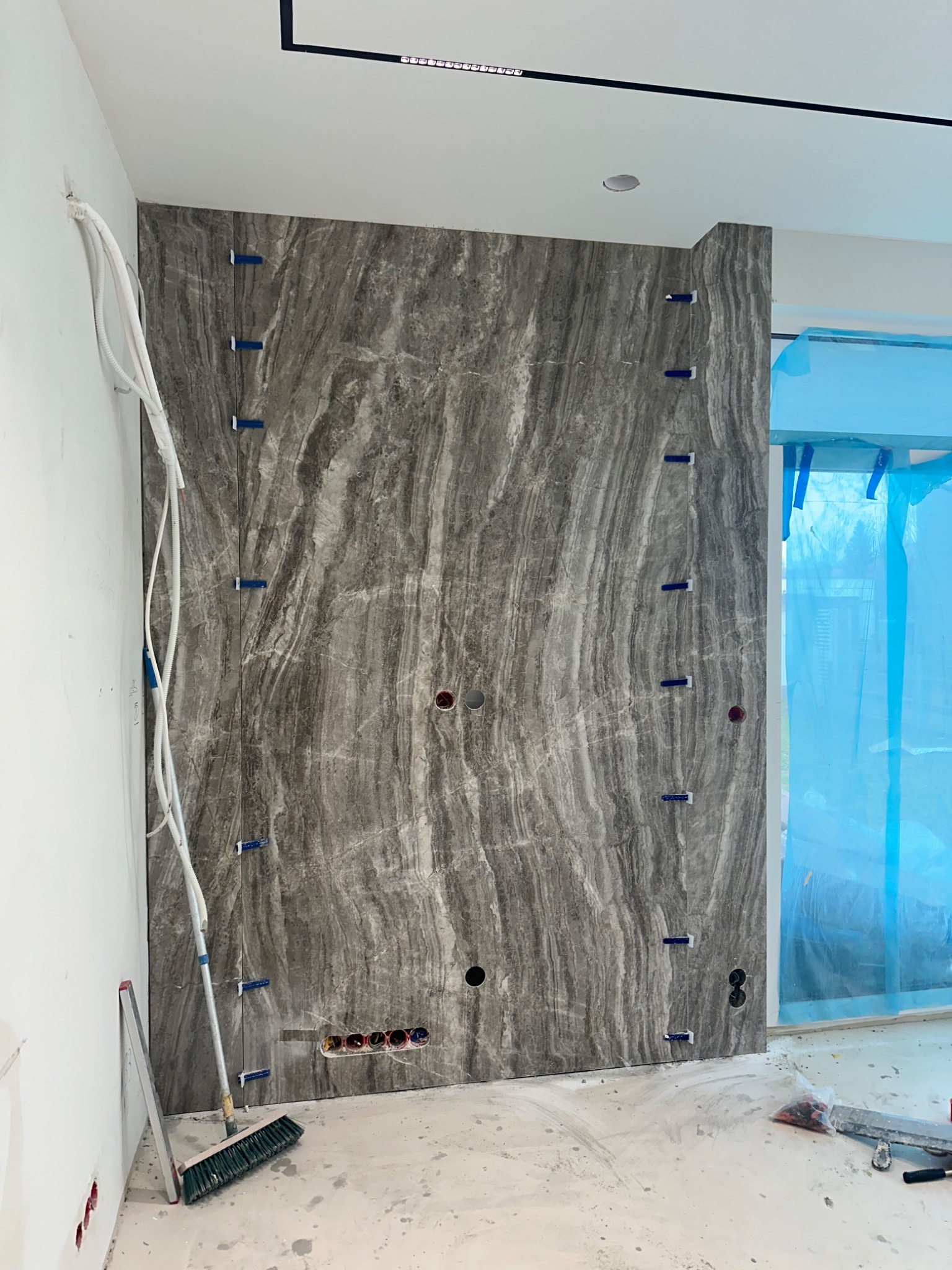 Ściana wyłożona płytami imitującymi marmur, z widocznymi otworami na gniazdka i niebieskimi klinami dystansowymi, w trakcie prac wykończeniowych w pomieszczeniu z zabezpieczonym oknem.