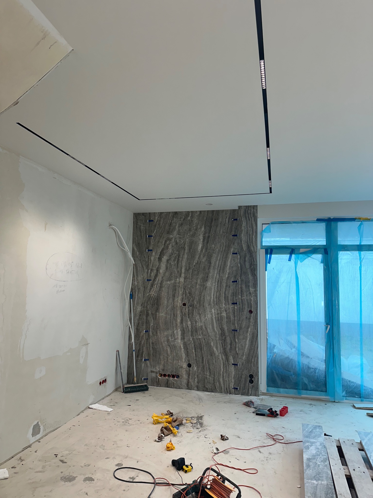 Wnętrze w trakcie prac wykończeniowych: ściana z szarej okładziny imitującej marmur, widoczne przewody elektryczne, listwy oświetleniowe na suficie, okno zabezpieczone folią ochronną i narzędzia...