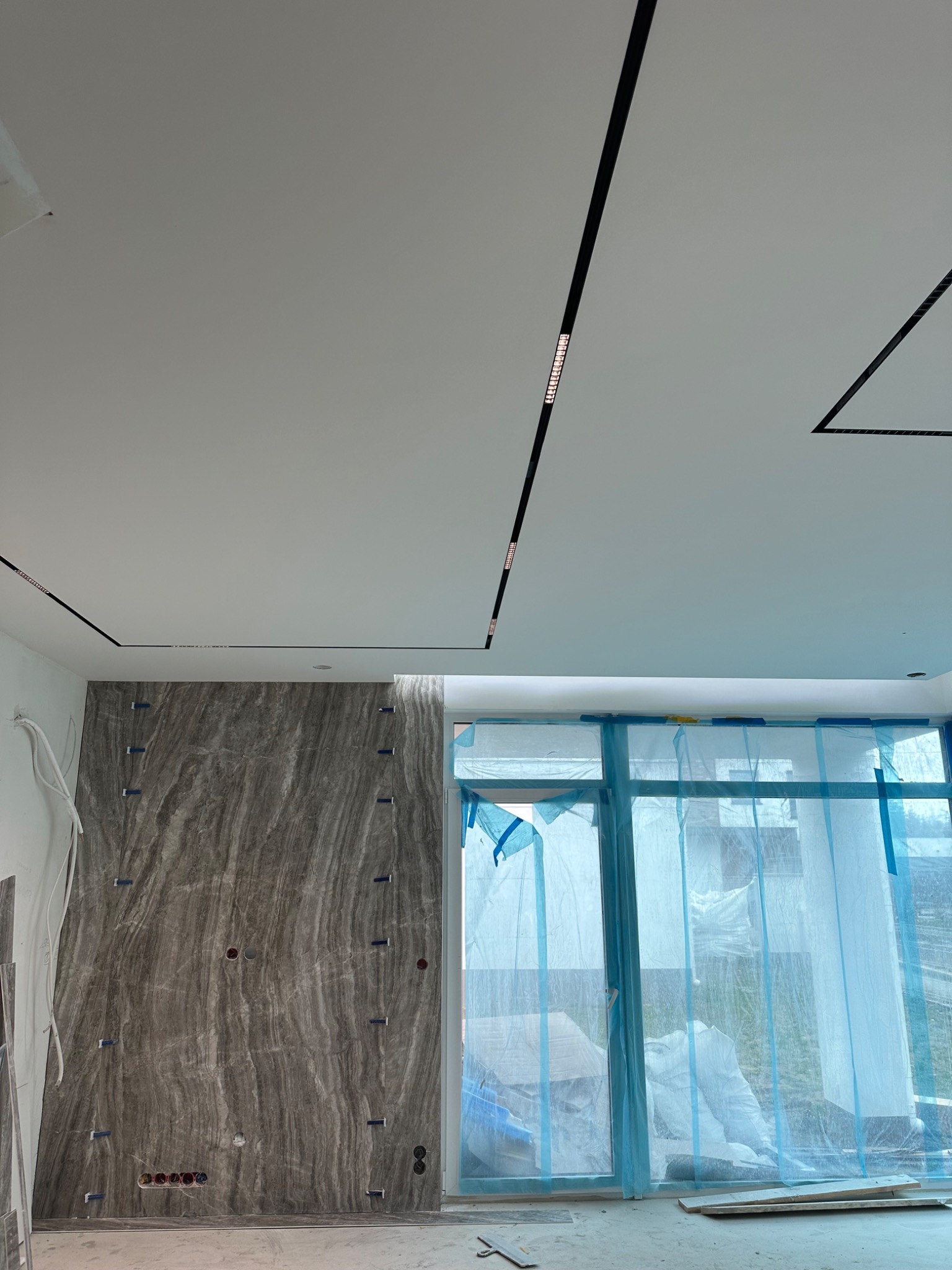 Wnętrze w trakcie wykańczania: ściana wyłożona płytami imitującymi marmur z widocznymi otworami na gniazdka elektryczne i niebieskimi klinami, sufit z podłużnym oświetleniem LED, okna zabezpieczone...