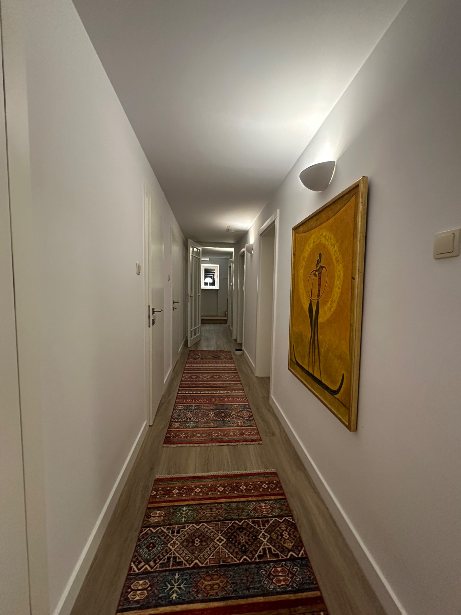 Długi, wąski korytarz z białymi ścianami, oświetlony kinkietami, z drewnopodobną podłogą i dwoma orientalnymi dywanami. Na ścianie wisi obraz w złotej ramie przedstawiający abstrakcyjną postać.