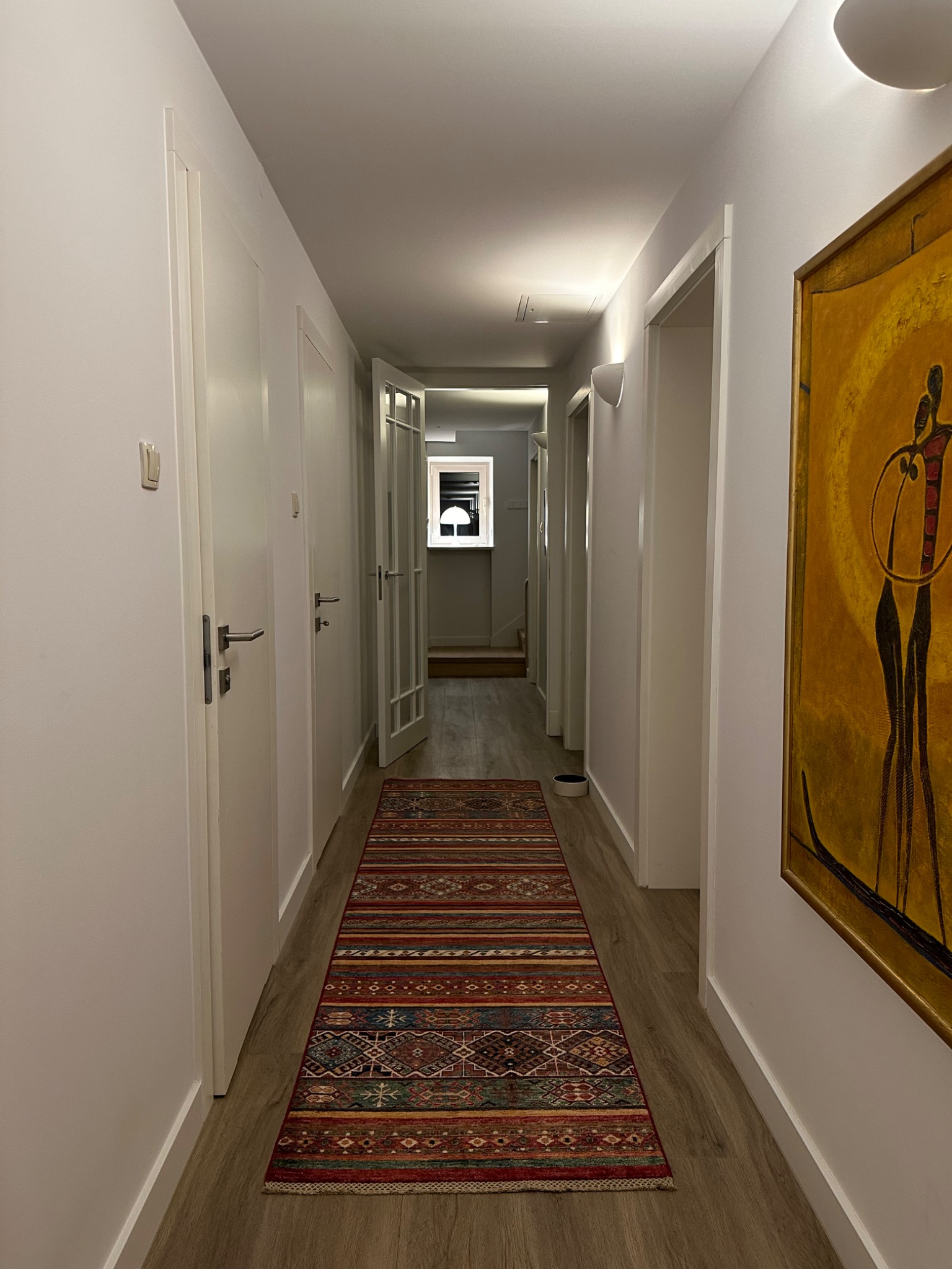 Długi, wąski korytarz z panelami podłogowymi i wzorzystym dywanem w odcieniach czerwieni i brązu, zakończony oświetlonym pomieszczeniem. Na ścianie wisi abstrakcyjny obraz w złotej ramie.