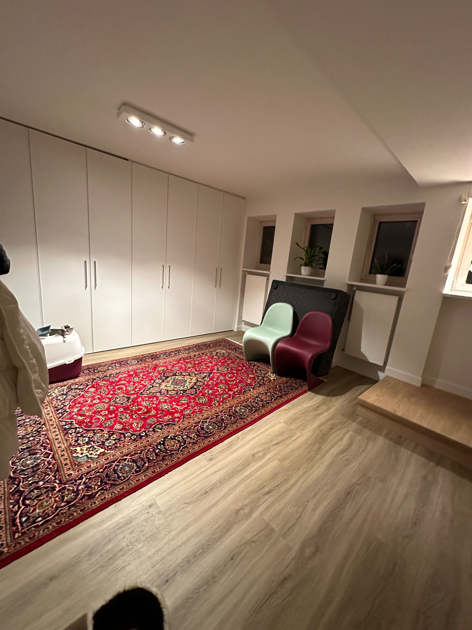 Jasne wnętrze z panelami podłogowymi, wzorzystym dywanem orientalnym, białymi szafami w zabudowie i dwoma krzesłami z tworzywa sztucznego w kolorach pastelowej zieleni i bordo.