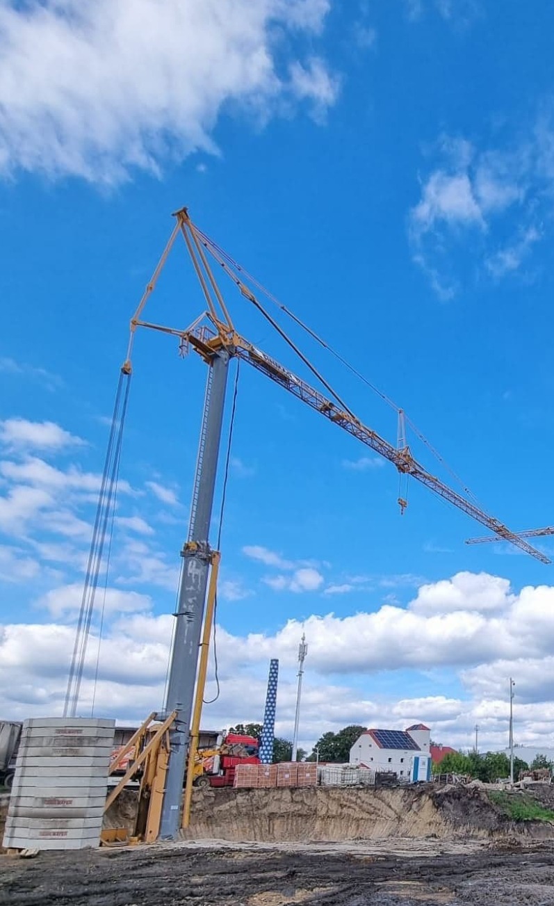 Żuraw wieżowy Potain HDT 80 na placu budowy z wykopem, widoczne betonowe płyty i cegły, w tle budynek z panelami słonecznymi i błękitne niebo z chmurami.